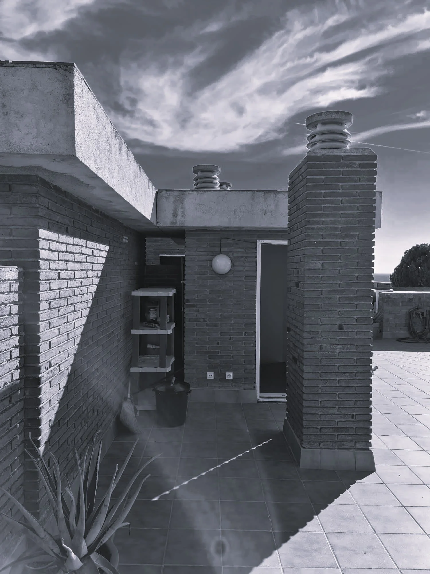 Outdoor rooftop area with brick walls, a small window, a door, a potted plant, a hanging lamp, and a vent on the roof under cloudy sky