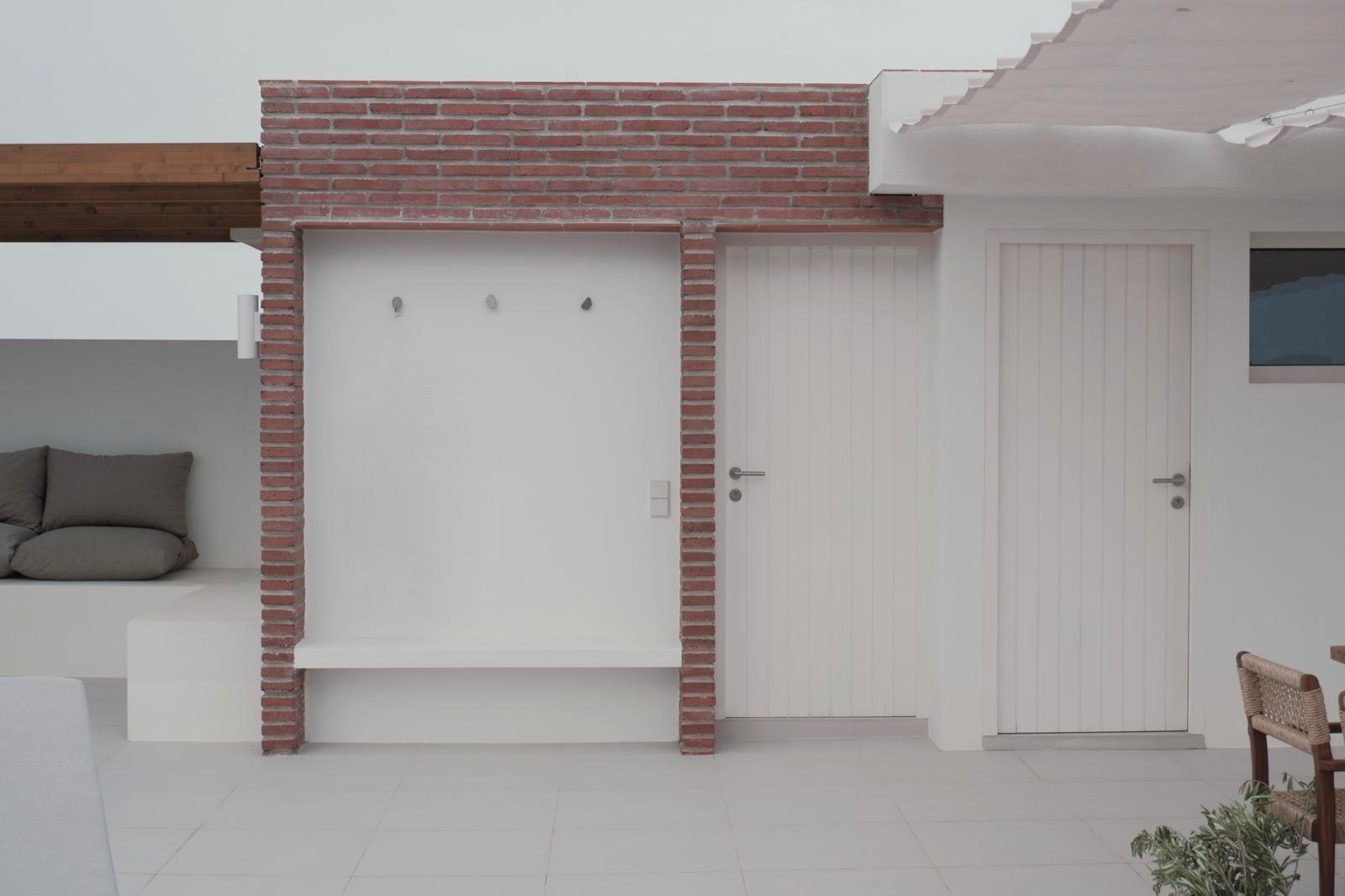 Interior of a modern minimalist room with white walls, a brick accent wall, and two white paneled doors. The left side has a built-in seating area with gray cushions, and the right side has a window and a brown woven chair. There are some small hooks or pegs on the wall.