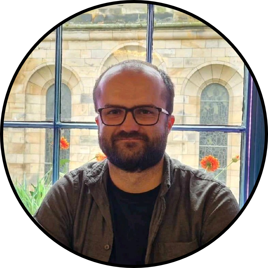 A man with glasses and a beard sitting in front of a large window with a stone building and flowers in the background.