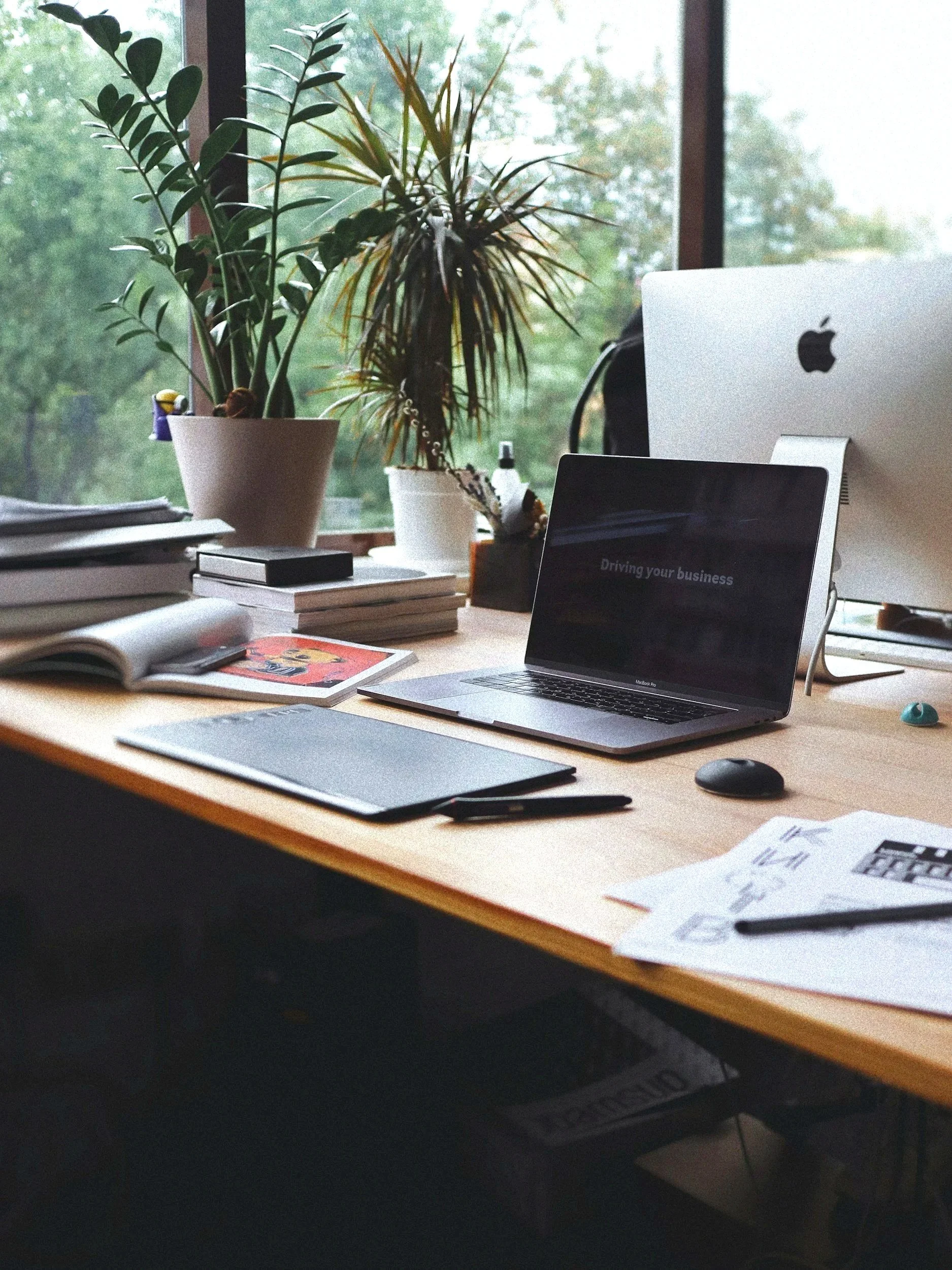 Organized office desk with two potted plants, various notebooks, papers, and pens, a laptop displaying 'Driving your business,' an iMac, and a computer mouse, with a large window showing greenery outside.