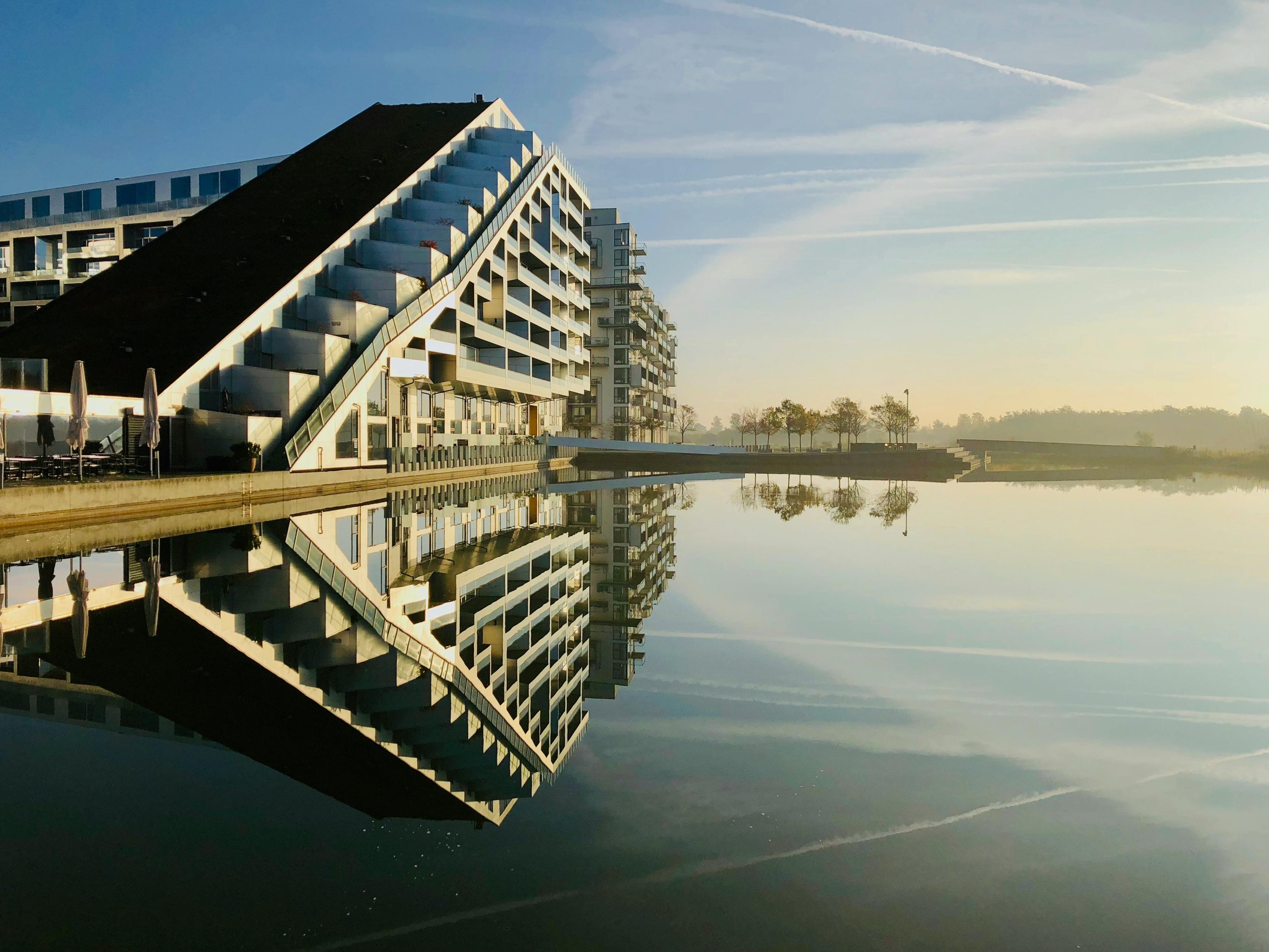 Modern multi-story residential building with balconies, situated beside a calm body of water, reflecting the building and a clear sky with some clouds.
