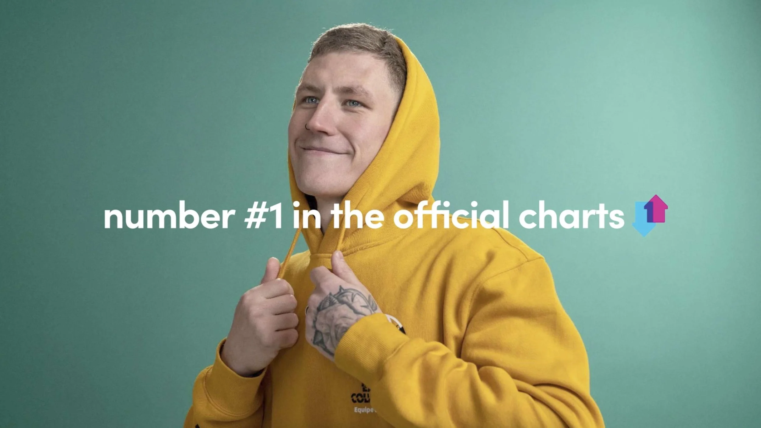 A young man wearing a yellow hoodie with a tattoo on his hand, smiling with a confident expression, standing against a green background.
