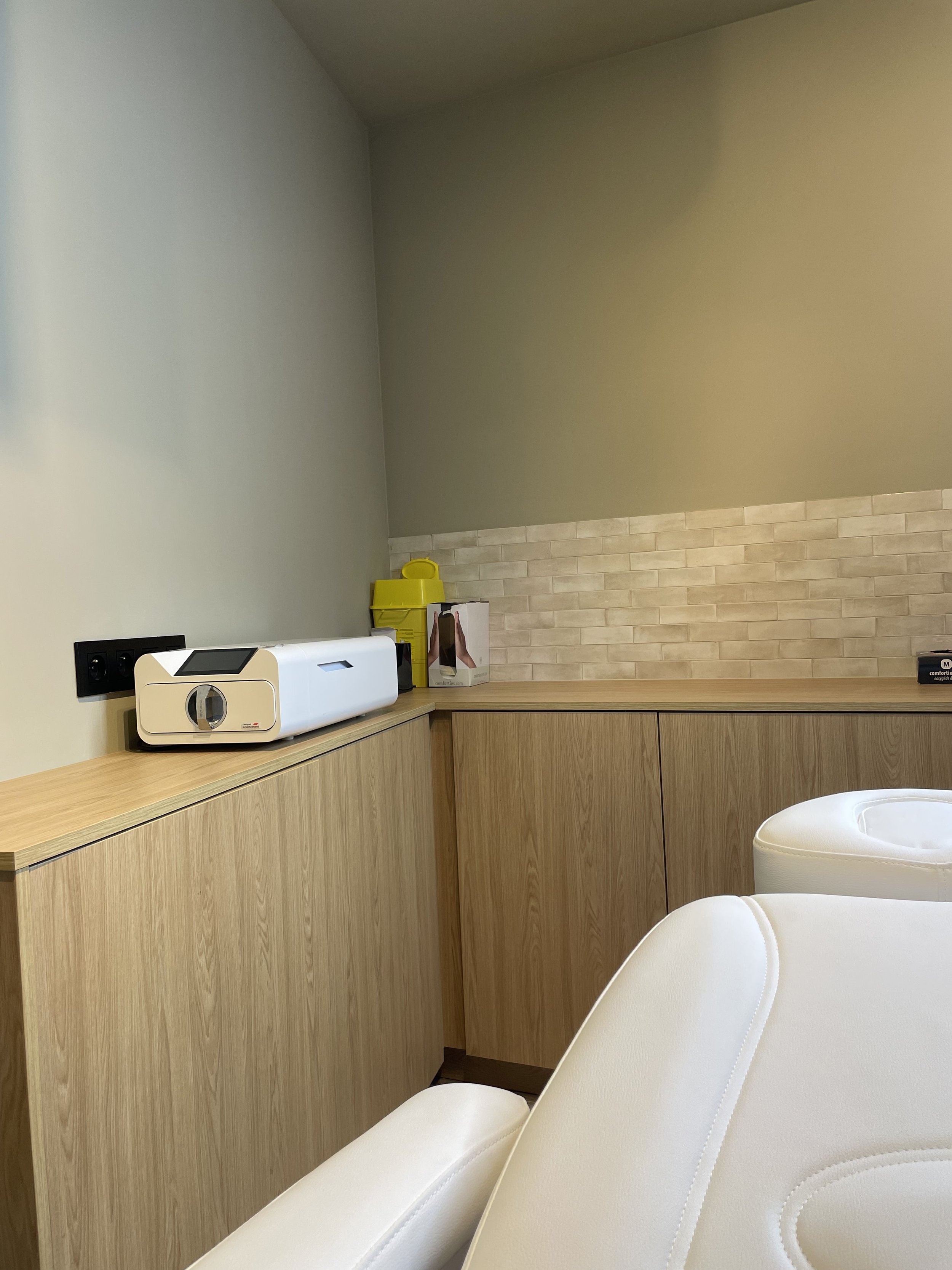 A corner of a salon or spa treatment room with white reclining chairs, a wooden cabinet, and a countertop with a white ultrasound or laser machine, yellow disinfectant or sanitizer bottles, and boxed products on top.