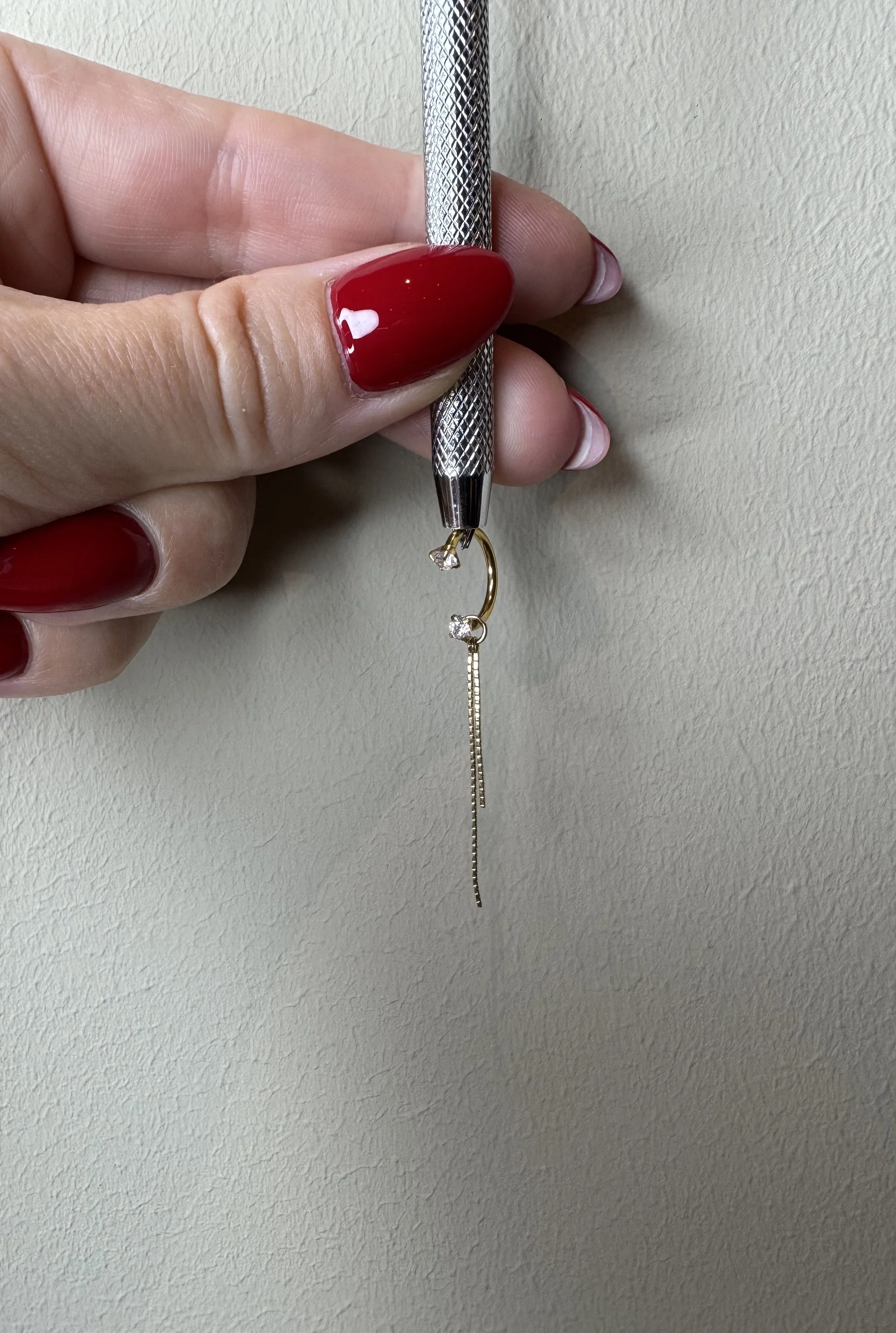 Close-up of a hand with red-painted nails holding a small drill, working on a gold earring with a dangling chains and diamonds, against a beige wall.