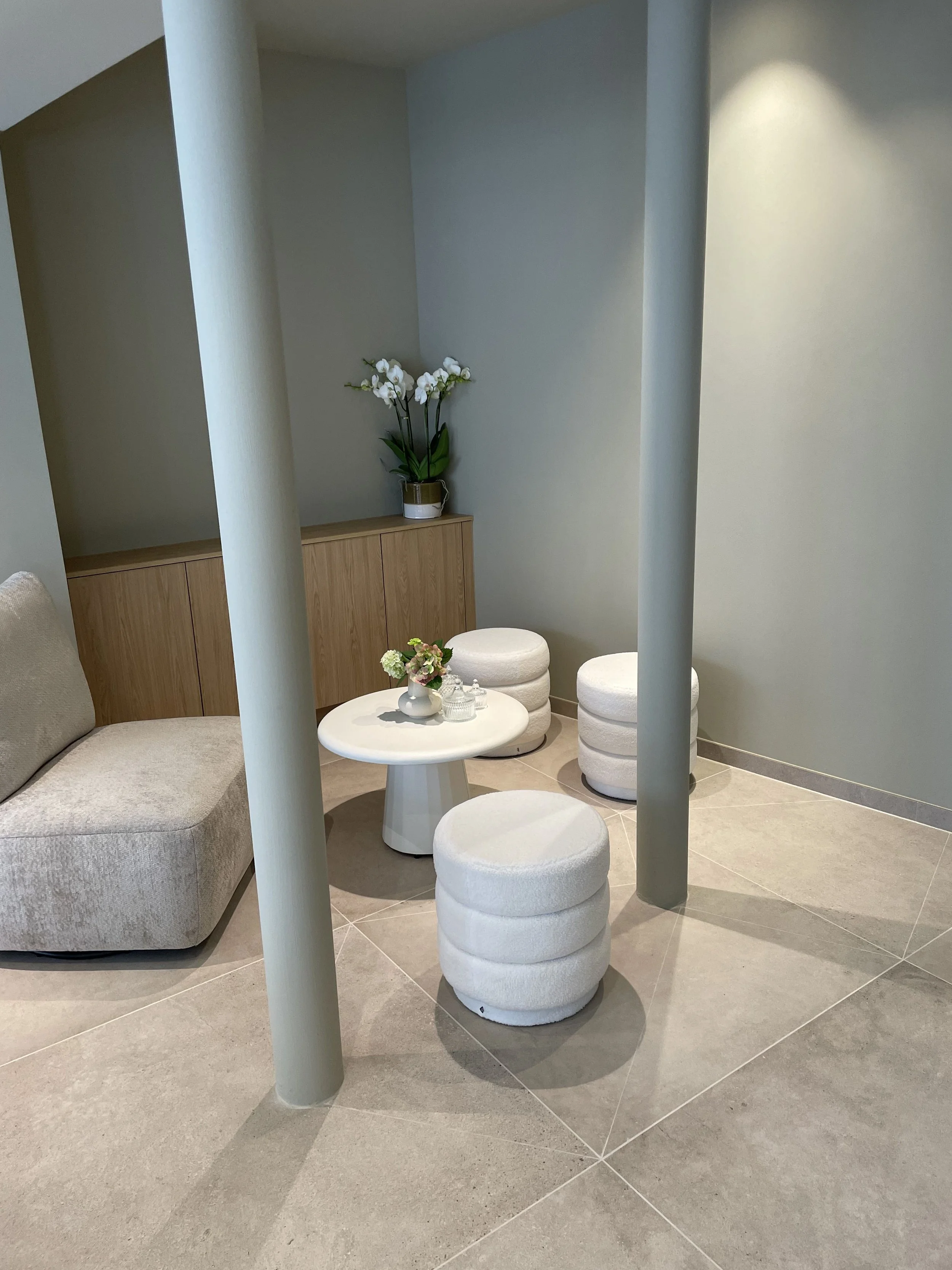 A cozy seating area with a beige curved sofa, a round white table with a flower arrangement, three white stacked cushioned stools, a wooden cabinet with a potted orchid, and a minimalist wall with soft lighting.