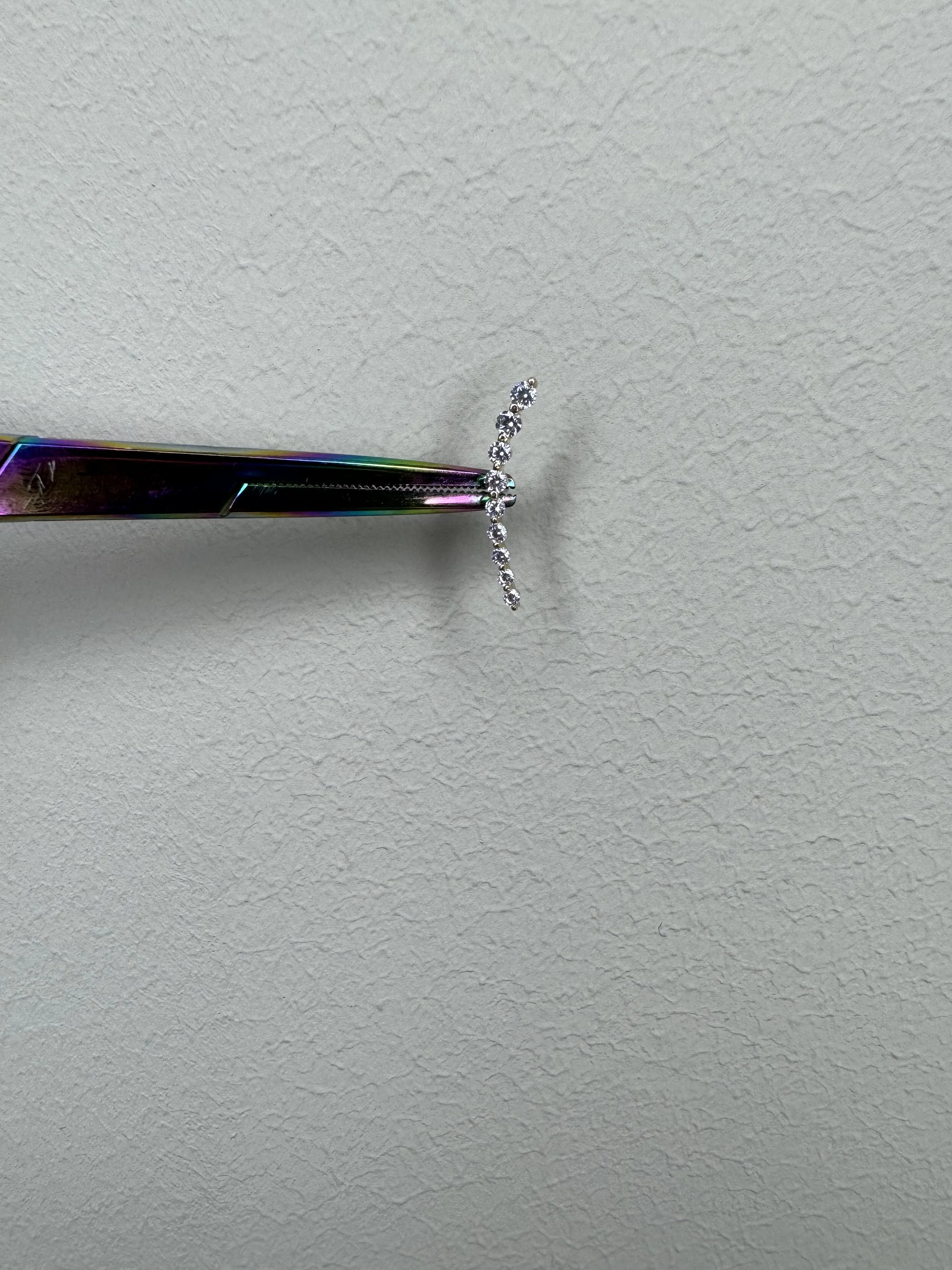 A close-up of a pair of tweezers holding a silver ring with small gemstones arranged in a curved pattern, against a plain textured background.