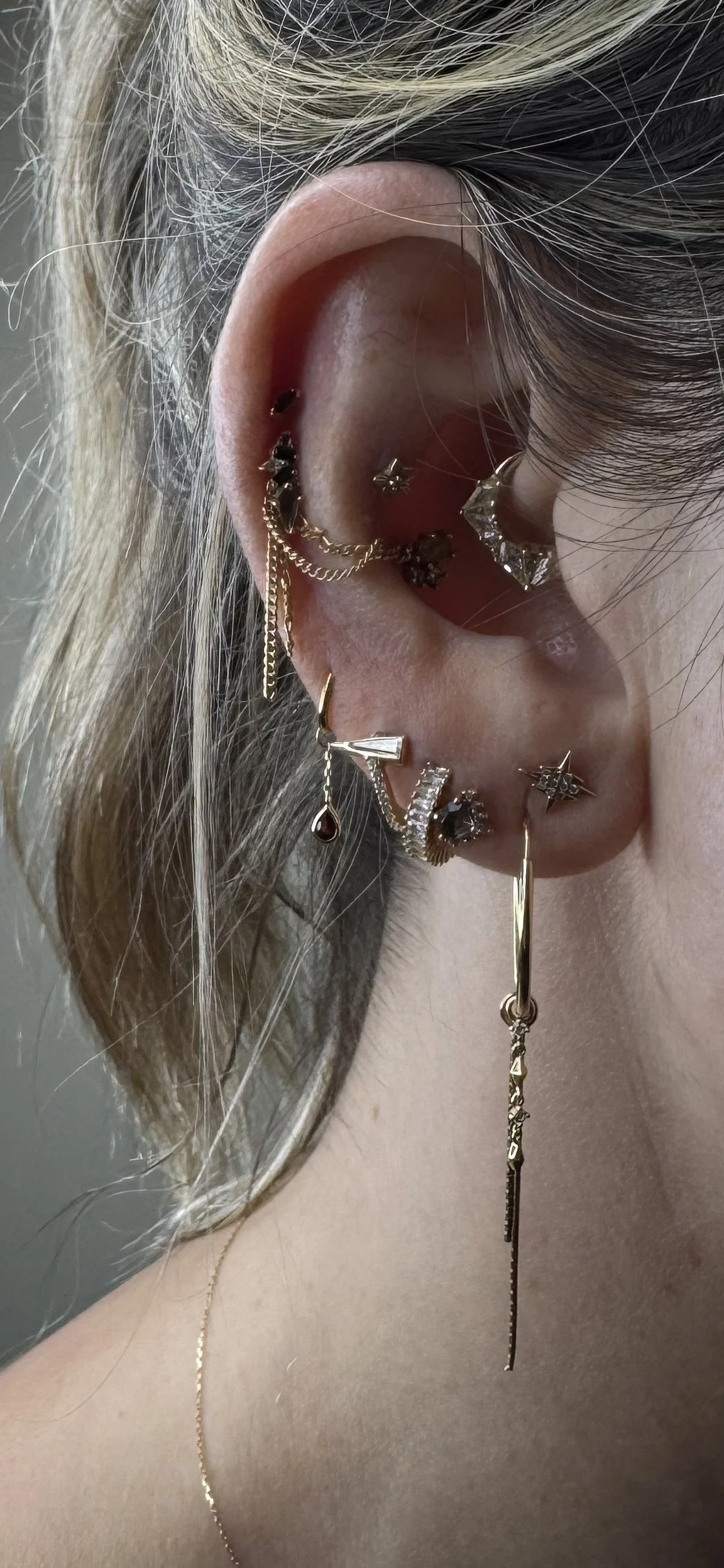 Close-up of a person's ear with multiple gold and silver earrings, including hoops, studs, and chains, some with gemstones or decorative elements.