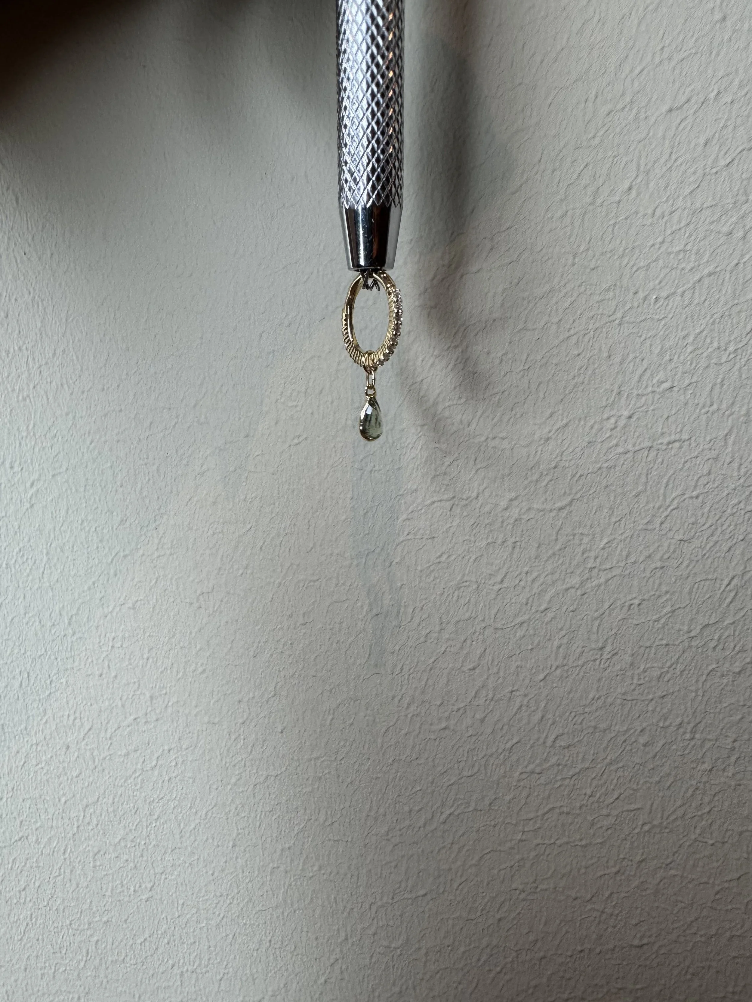 Small gold ring with a dangling teardrop-shaped gemstone, hanging from a metal textured tool.