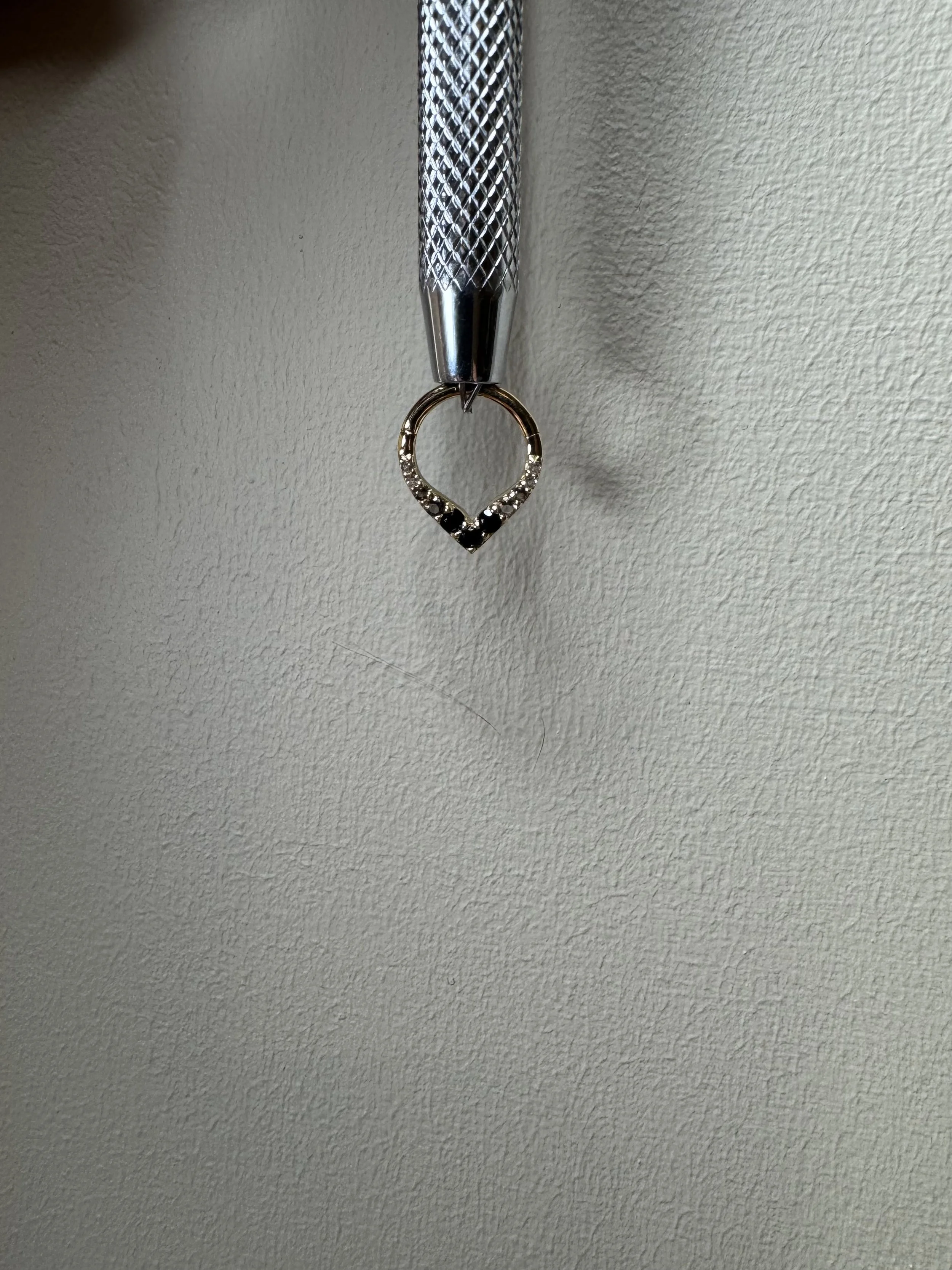 A close-up photo of a metallic ring with small gemstones, hanging from a textured metal tool against a plain wall background.