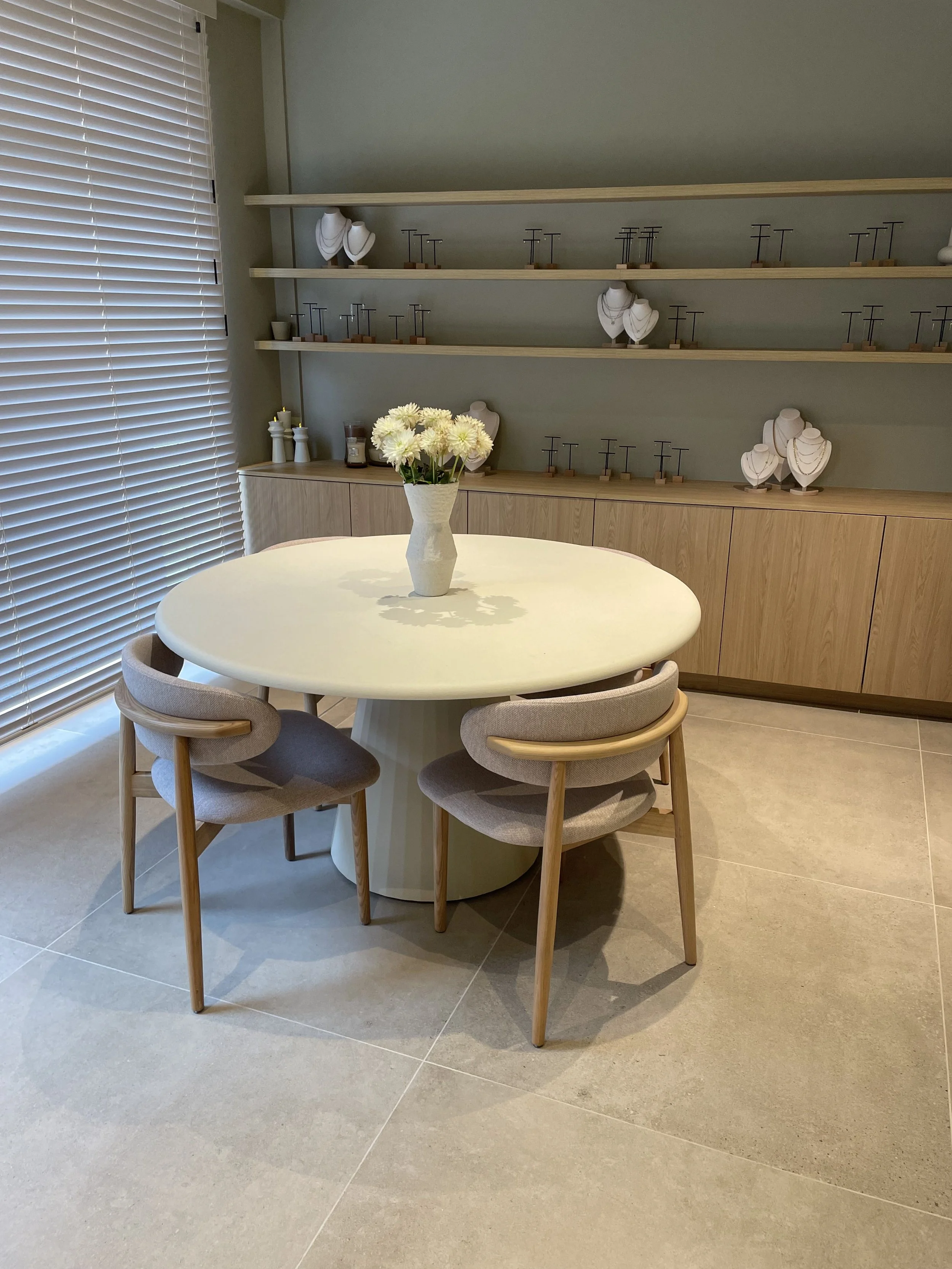 A round table with a beige vase of white flowers on top, surrounded by four beige upholstered chairs with wooden legs, next to a window with blinds, and a wooden sideboard with shelves displaying jewelry stands and decorative items.