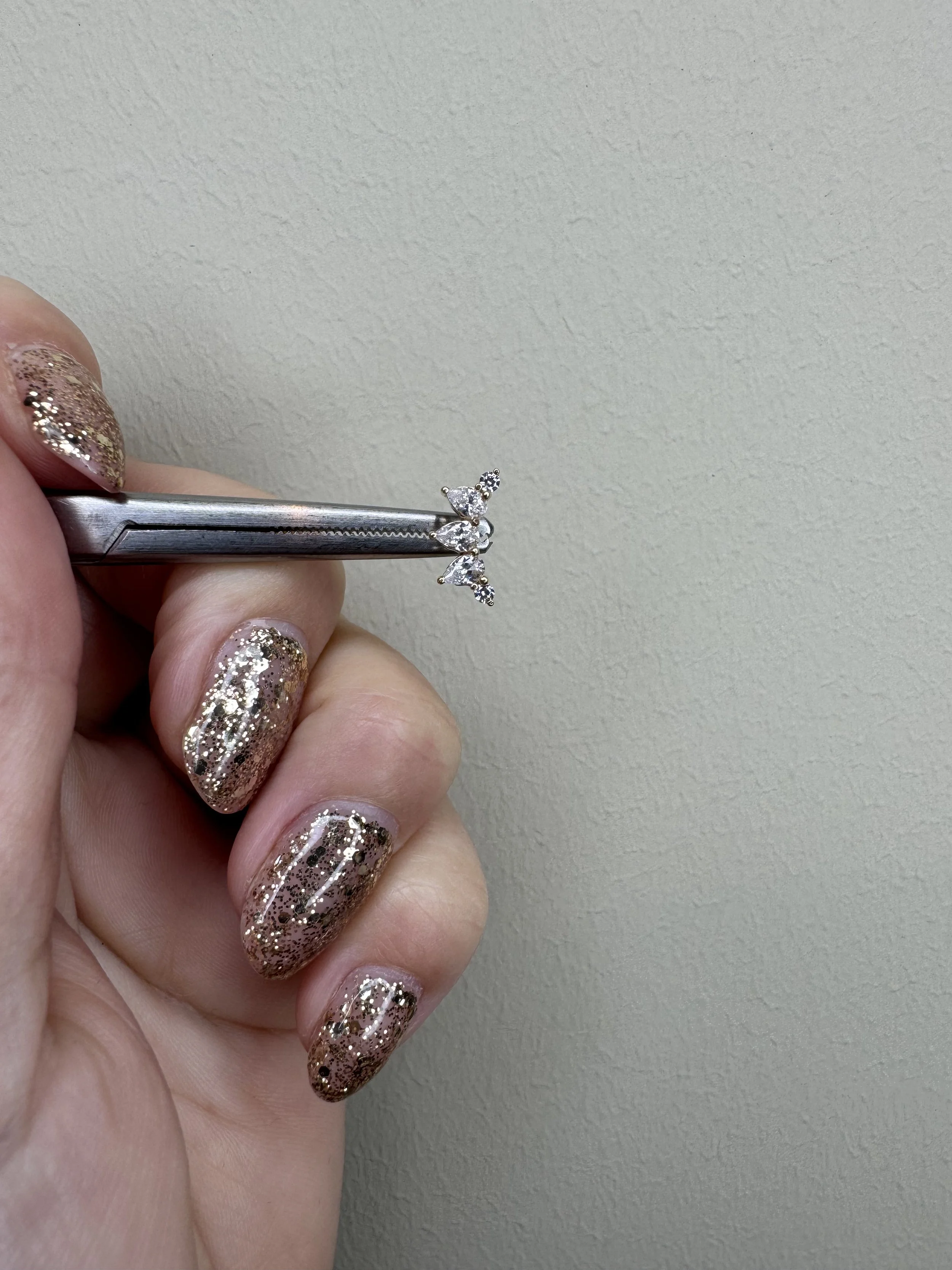 Hand holding a pair of tweezers with a small diamond ring against a plain wall background.