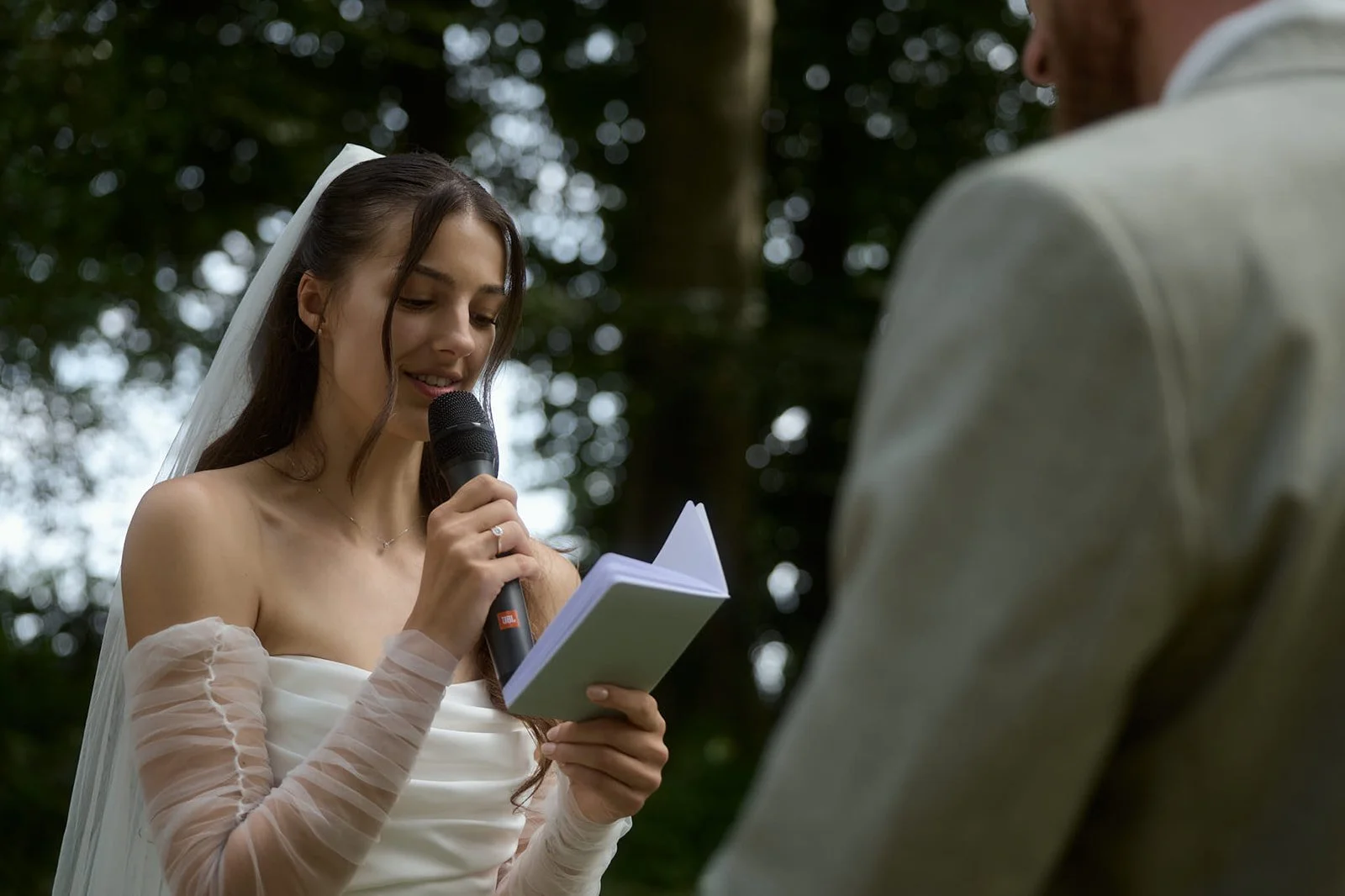 A young woman in a wedding dress is reading vows from a small notebook during an outdoor wedding ceremony, with a man partially visible in the foreground.