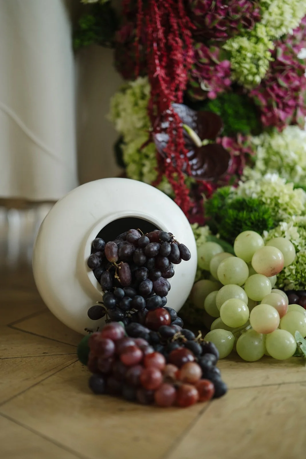 A white vase with black grapes spilling out onto a wooden surface, with various green and purple flowers in the background.
