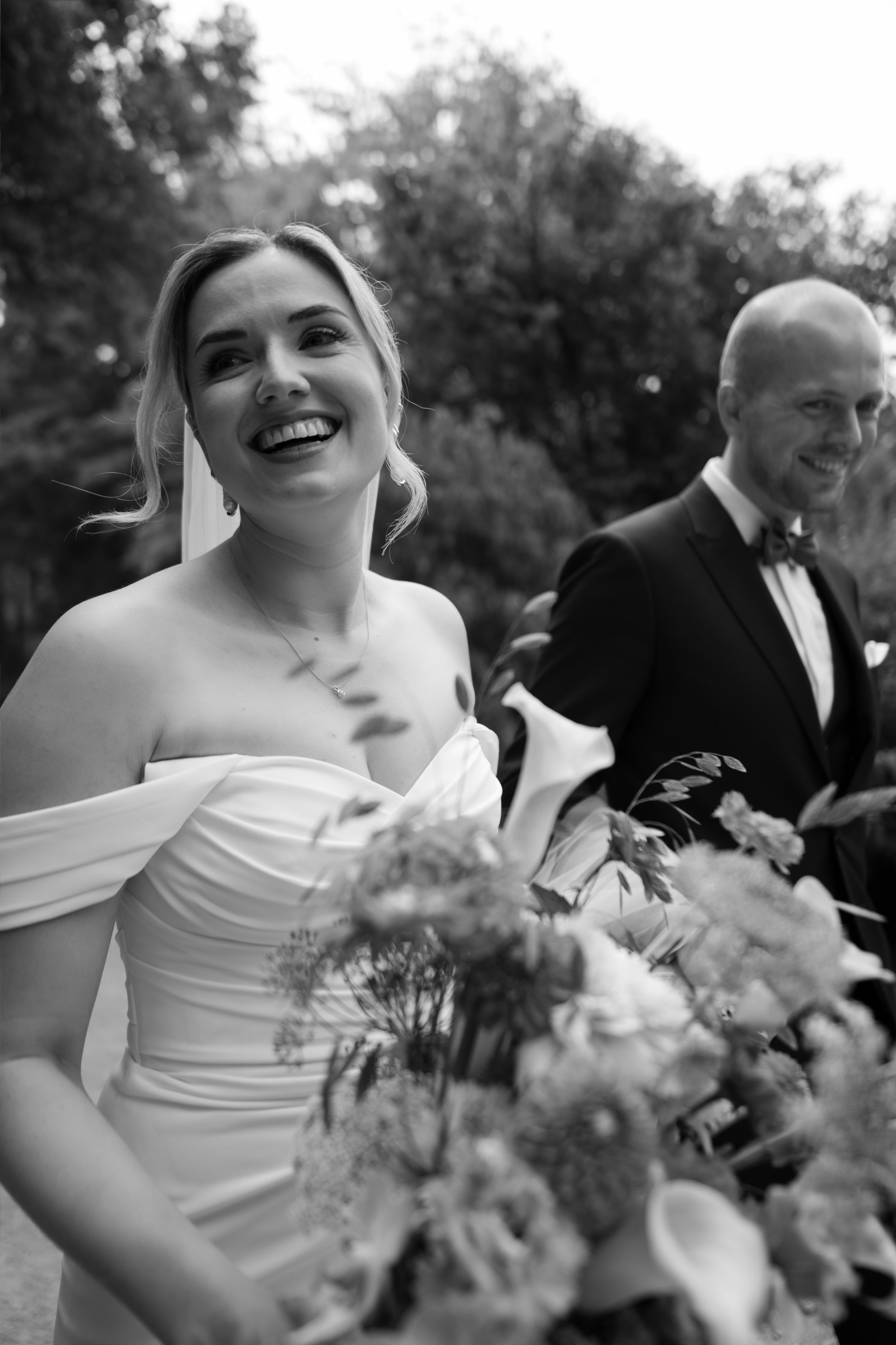 Black and white photo of a smiling woman in a wedding dress holding a bouquet, standing outdoors with trees in the background, and a man in a tuxedo smiling beside her.