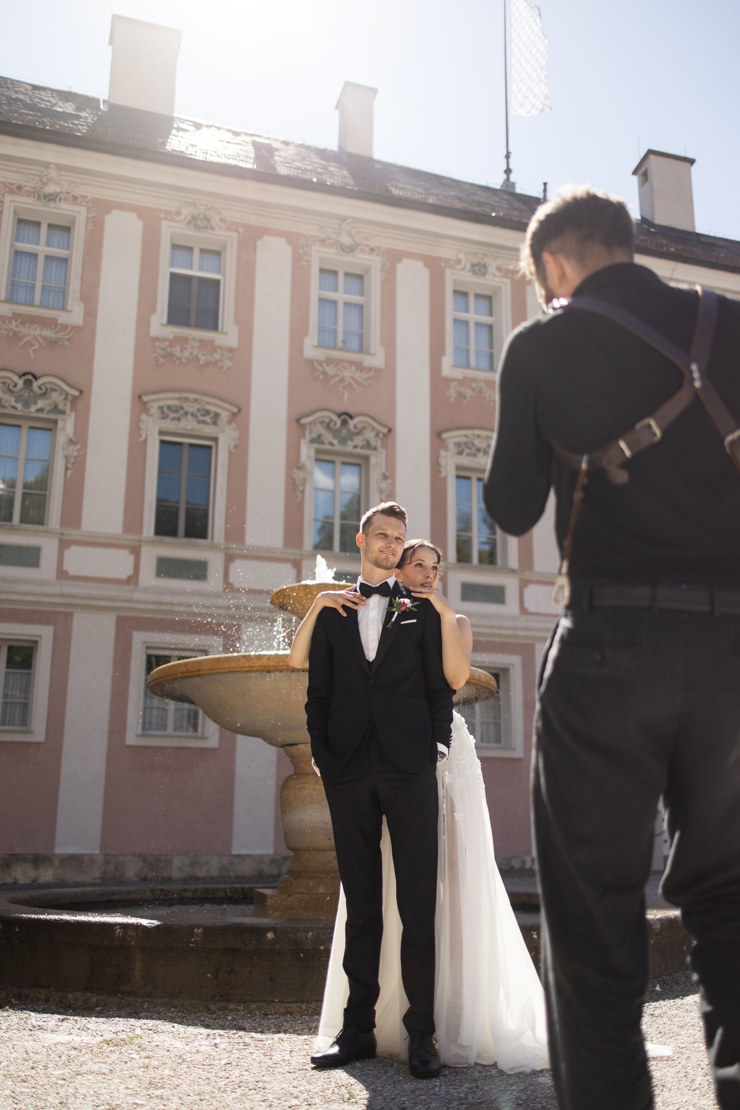 A bride and groom pose in front of a fountain and a pink ornate building, with a man taking a photo of them.
