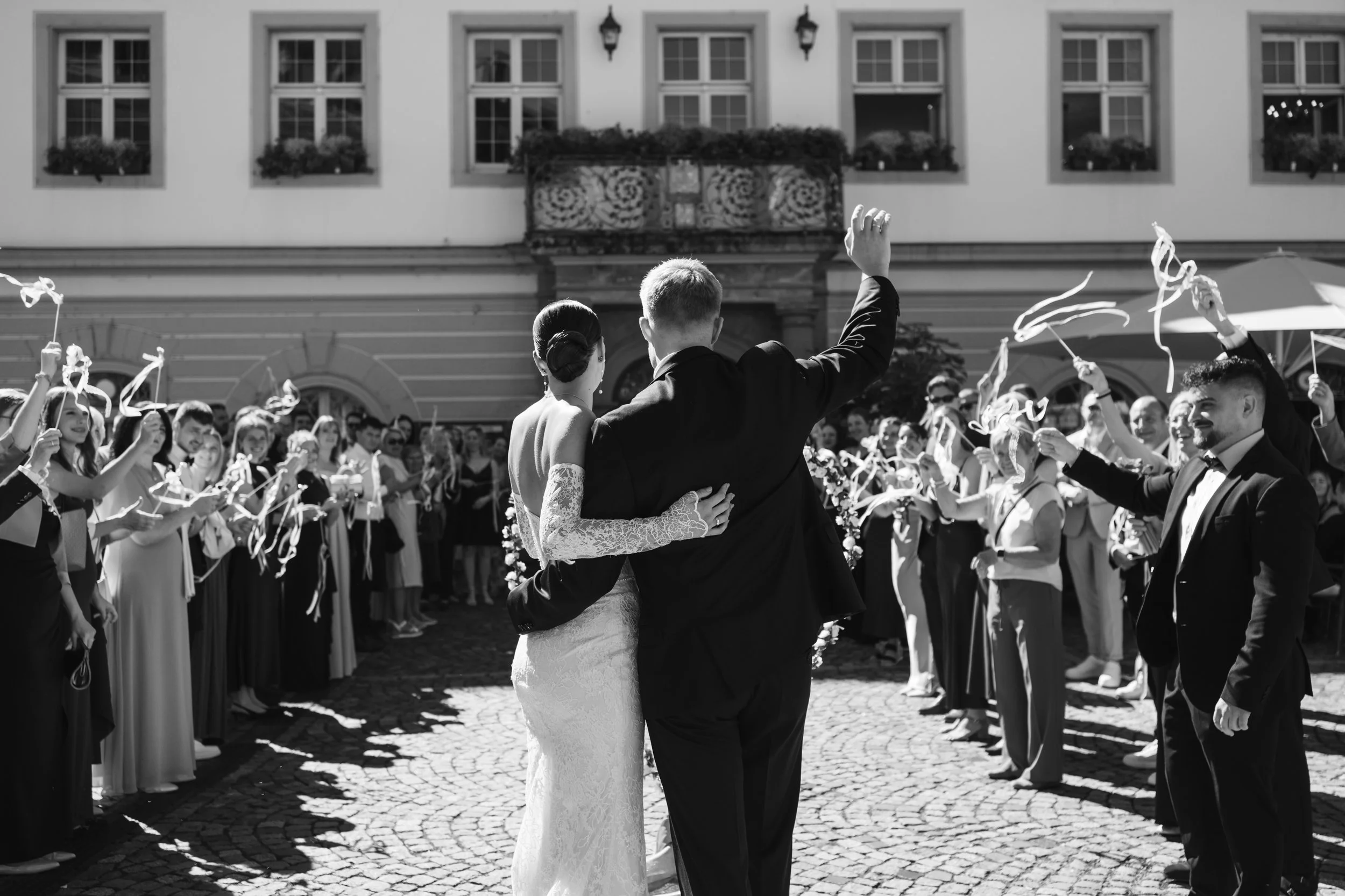 A black and white photo of a wedding scene showing a bride and groom dancing closely in the center, surrounded by guests holding streamers and celebrating outdoors in front of a historic building with multiple windows and flower boxes.