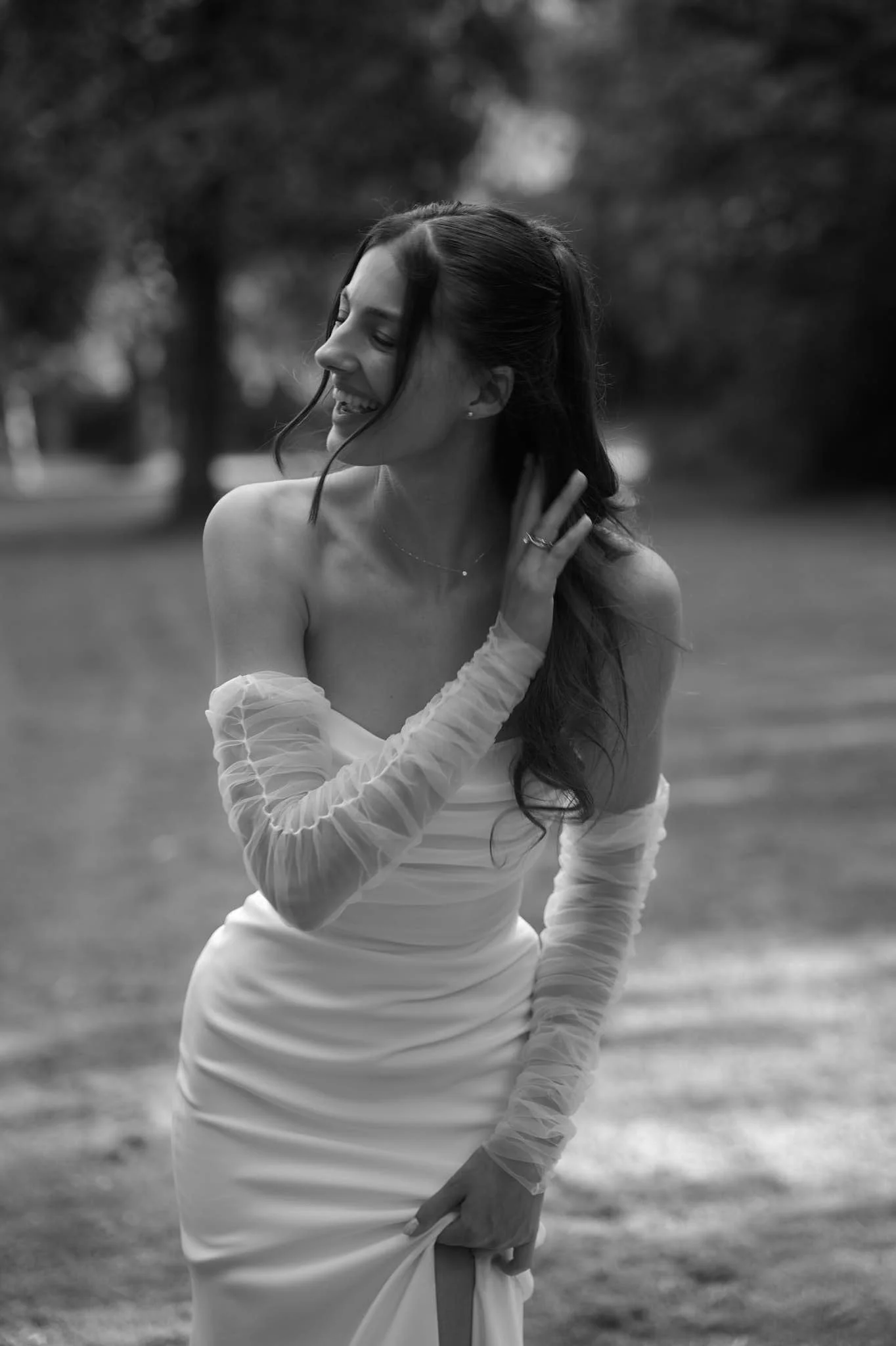 A woman in a strapless dress with sheer, ruffled long sleeves is outdoors, looking away and smiling with her eyes closed. She is touching her neck and holding the bottom of her dress with her other hand, in a natural, joyful pose.