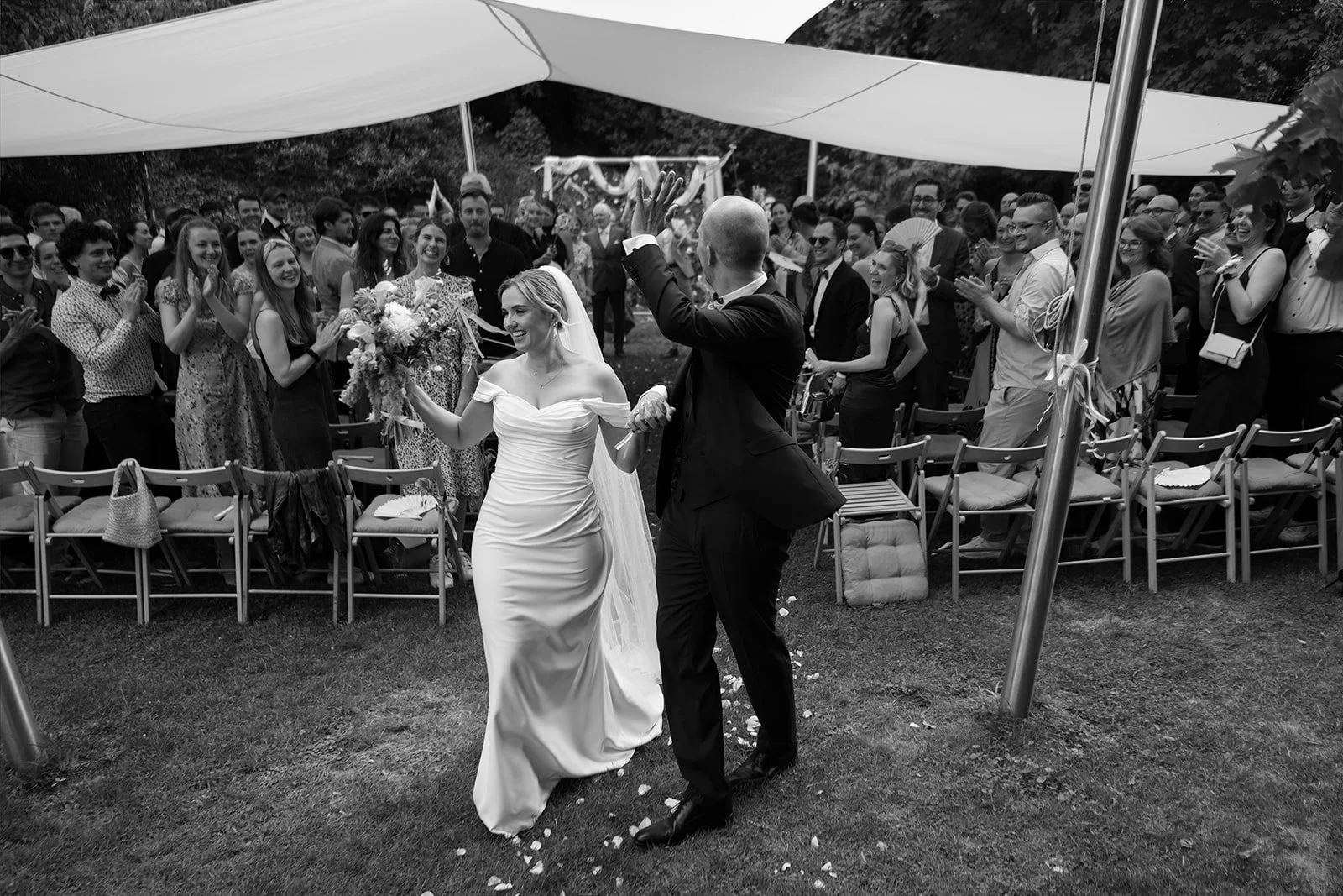 Black and white photo of a wedding ceremony outdoors. A bride in a white wedding gown and veil is holding a bouquet, smiling, and walking with a groom in a suit. Guests are clapping and cheering in the background, seated and standing under a canopy with trees in the distance.