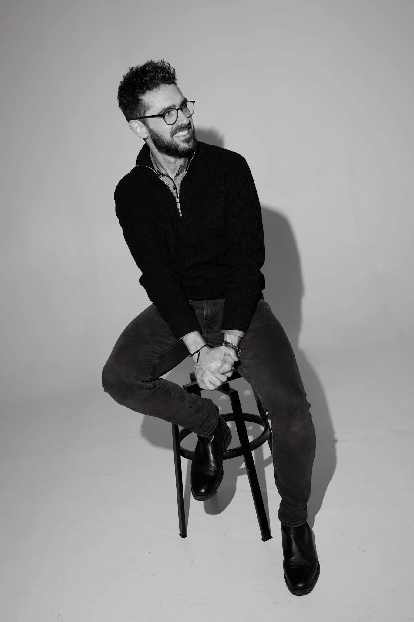 Black-and-white photo of a young man with curly hair, beard, glasses, and a smile, sitting on a high stool against a plain background. He is wearing a zip-up sweater, jeans, and boots, with his hands clasped in front of him.
