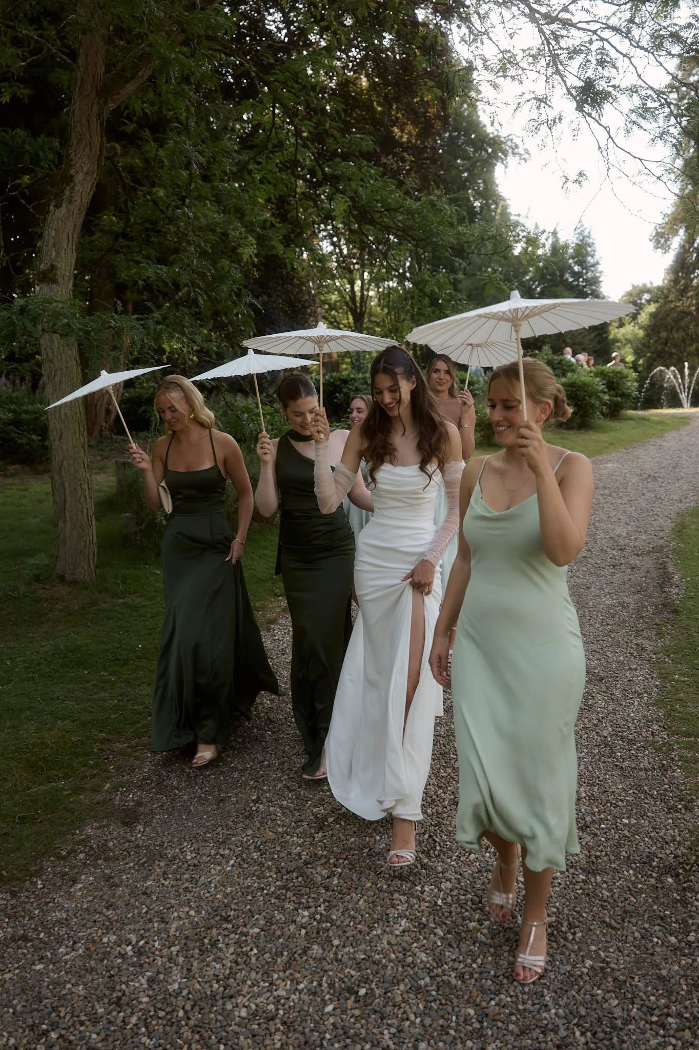 A group of women in elegant dresses walking outdoors in a garden, holding white parasols.