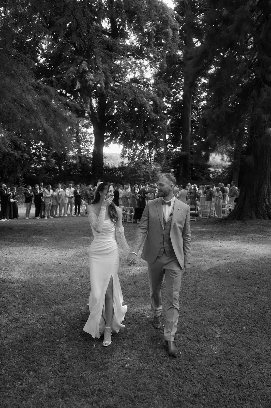 A black and white photograph of a newlywed couple holding hands and walking outdoors during their wedding ceremony. The bride is wearing a long, elegant dress with a slit, and the groom is wearing a suit with a bow tie. In the background, a group of 