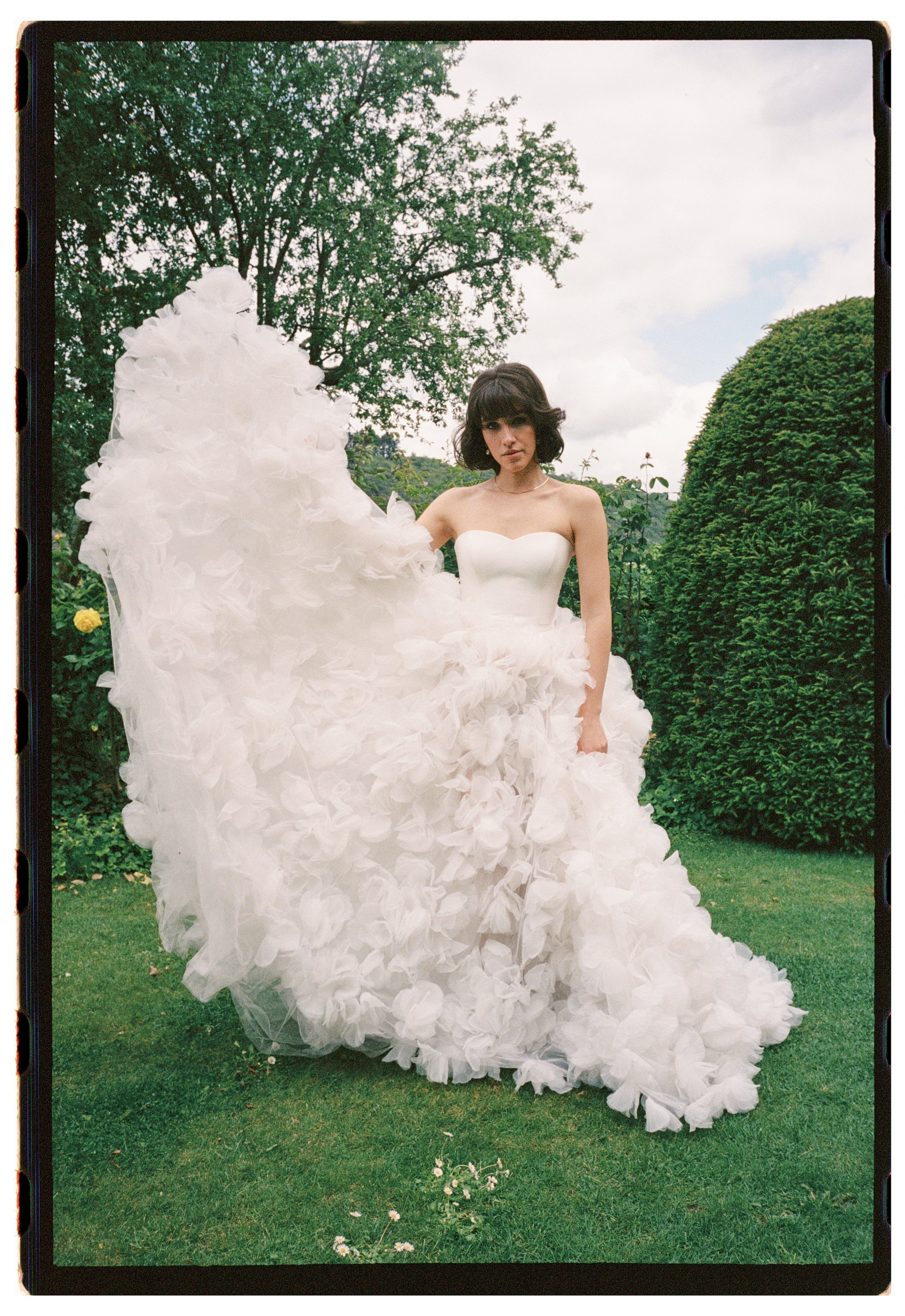 A woman in a strapless white wedding gown with a ruffled skirt standing in a garden with green trees and grass, holding up her dress.