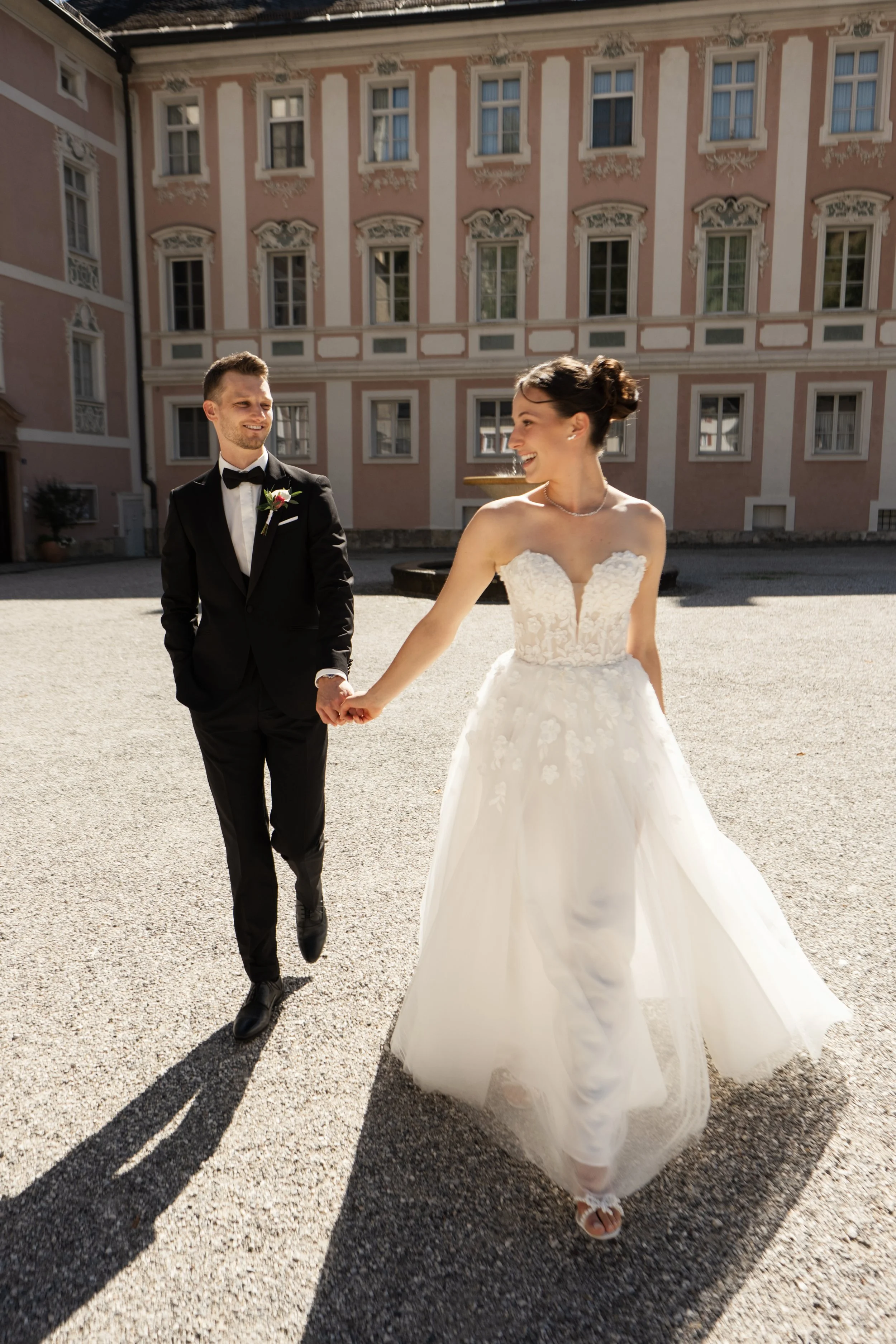 A bride and groom holding hands outdoors in front of a historic building, with the bride wearing a white wedding gown and the groom in a black tuxedo.
