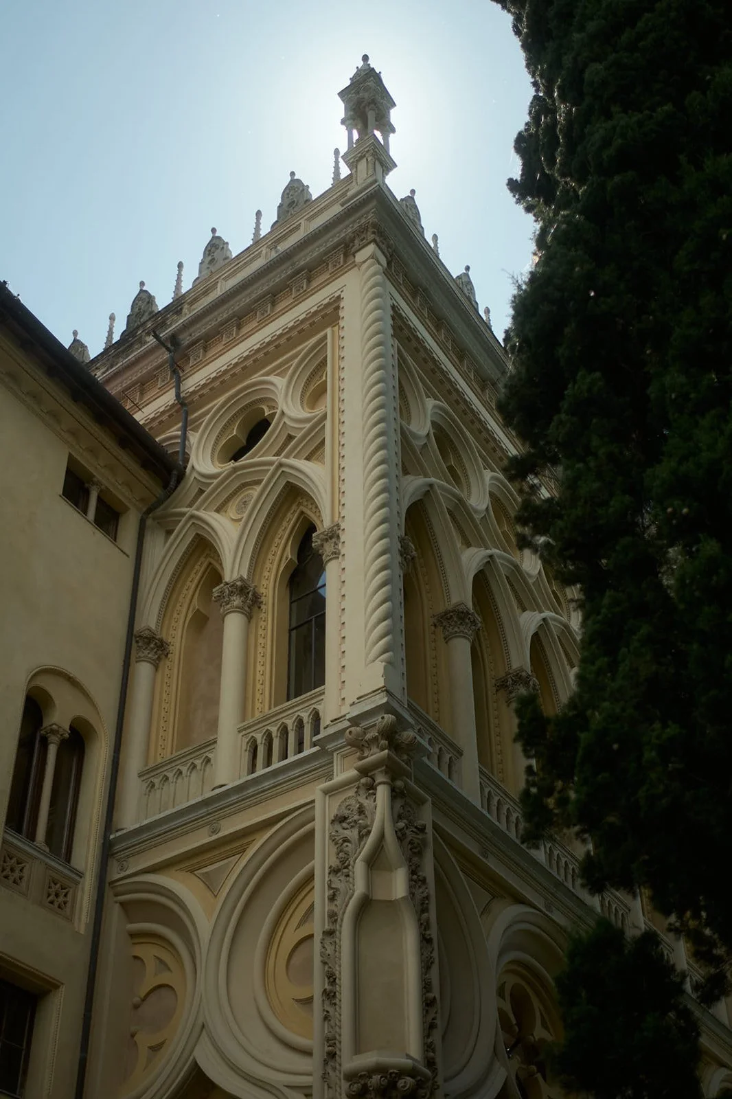 The ornate facade of a historic building, showcasing detailed architectural elements, arched windows, decorative columns, and a tower with statues on top, set against a clear sky with sunlight.