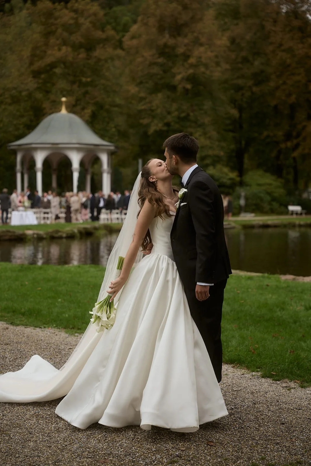 Bride and groom sharing a kiss outdoors during a wedding reception near a gazebo and a pond, with guests in the background.