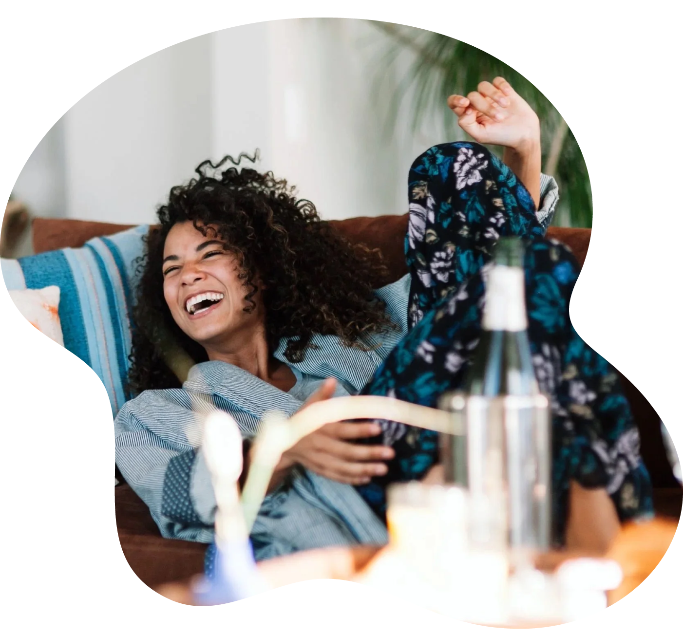 Woman with curly hair laughing and lying on a couch, wearing pajamas with a floral pattern, with a bottle and blurred objects in the foreground.