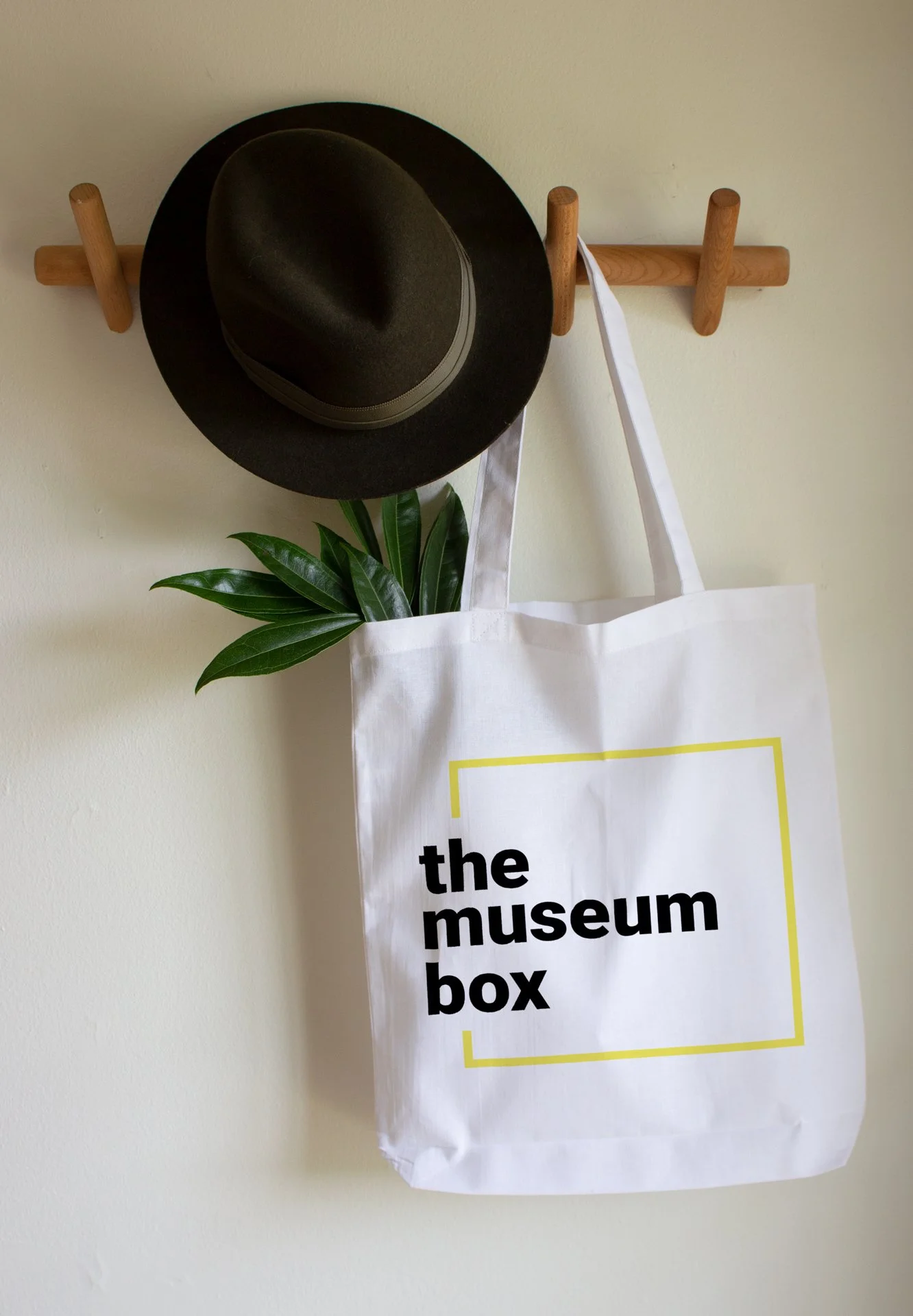 A mockup of a tote bag hanging on a wall hook featuring a "The Museum Box" logo.