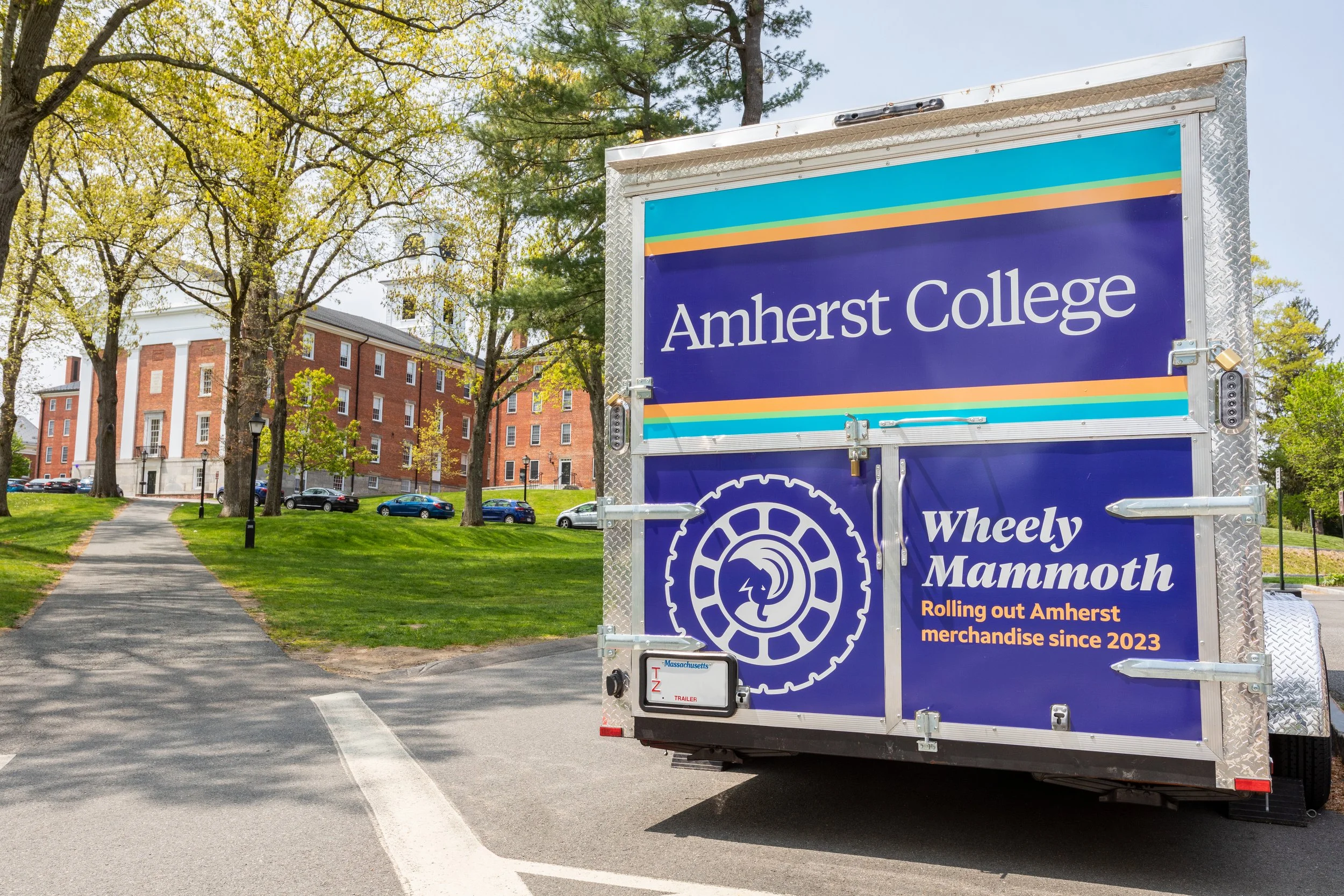 A rear view of a trailer with colorful stripes that reads "Amherst College" and then "Wheely Mammoth: Rolling out Amherst merchandise since 2023."