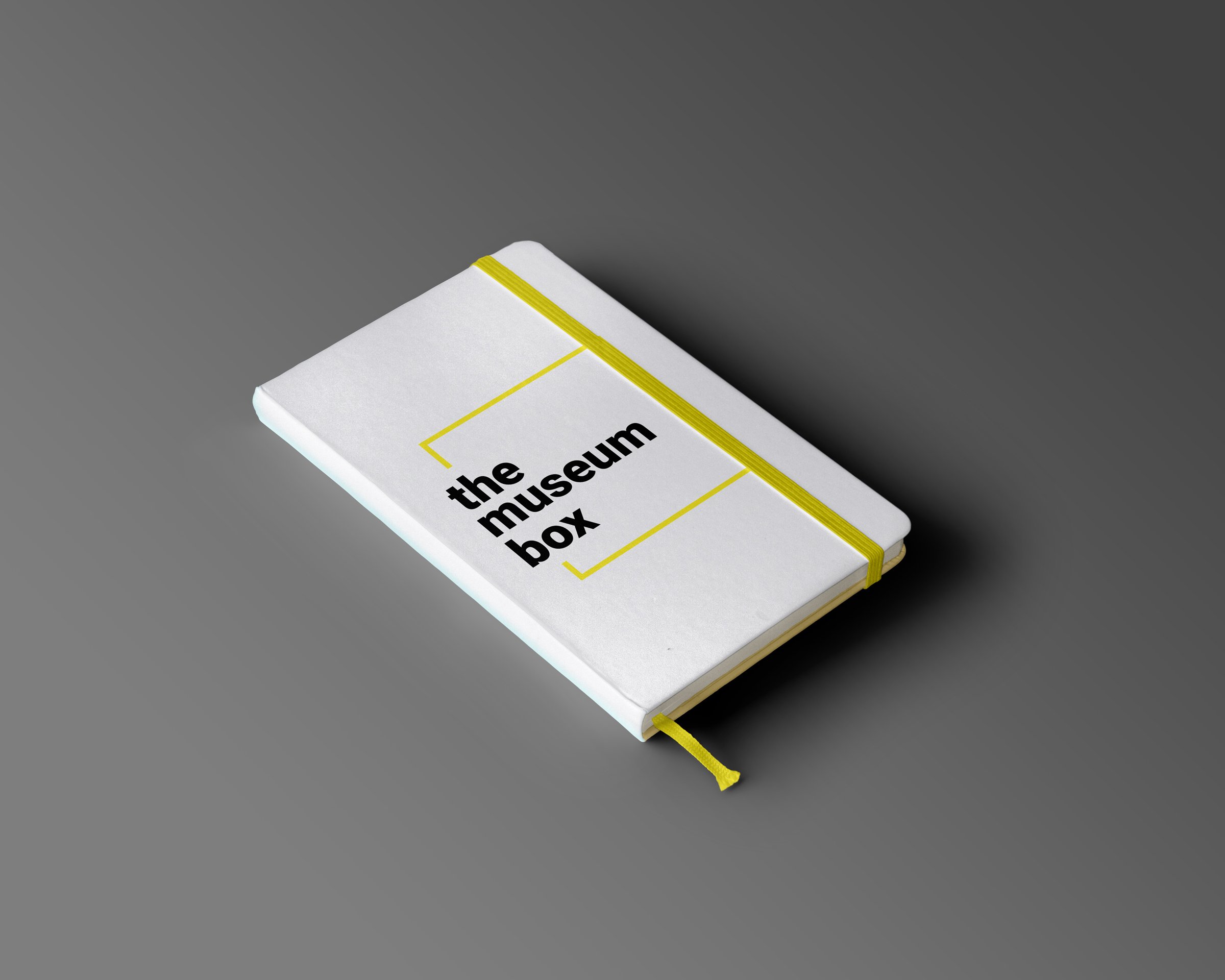 A mockup of a white notebook featuring a "The Museum Box" logo with a yellow ribbon and yellow bookmark.