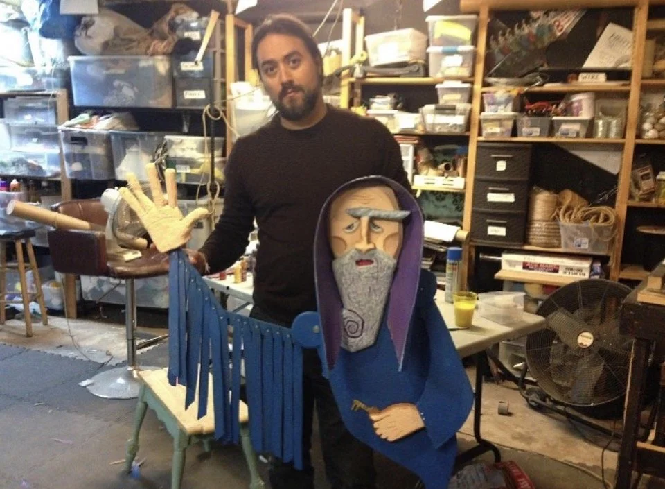 A man with a beard holding a puppet of an elderly wizard with a long gray beard, a purple hood, and a staff with a hand-shaped top in a cluttered workshop.