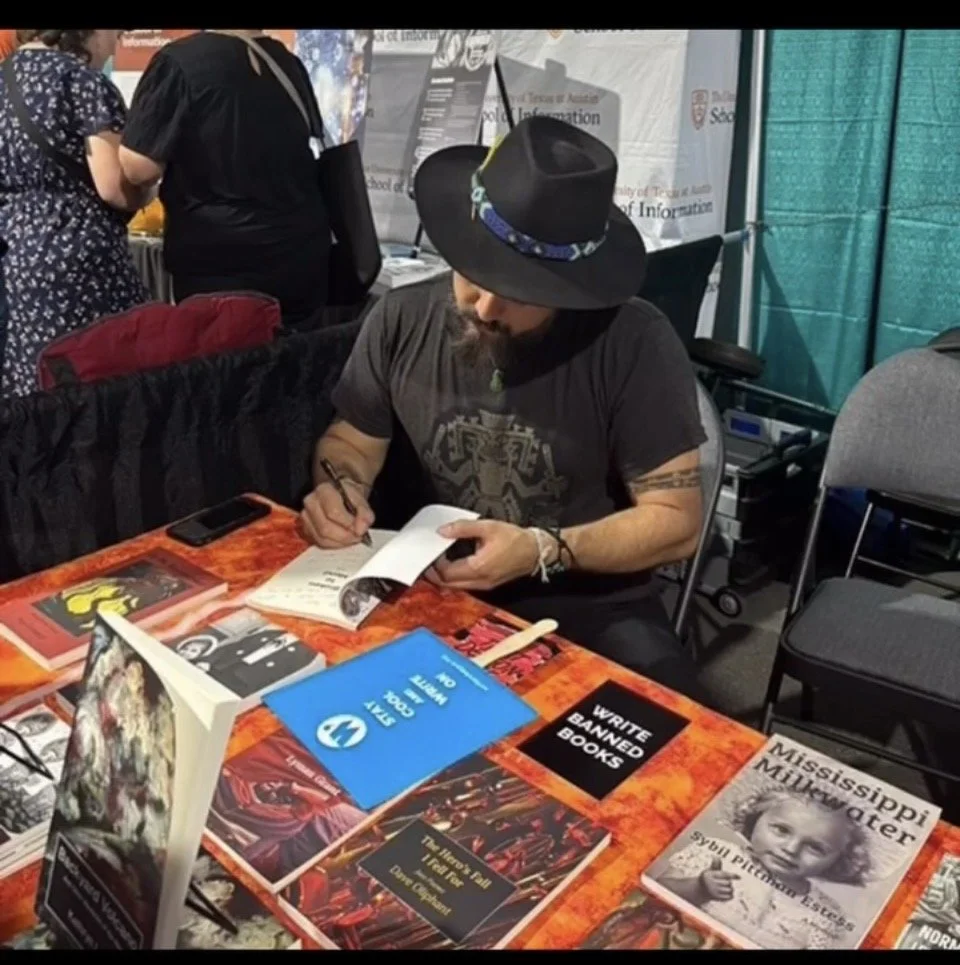 A man with a black hat and dark beard sitting at a table signing a book during a book signing event, with various books and promotional materials on the table.