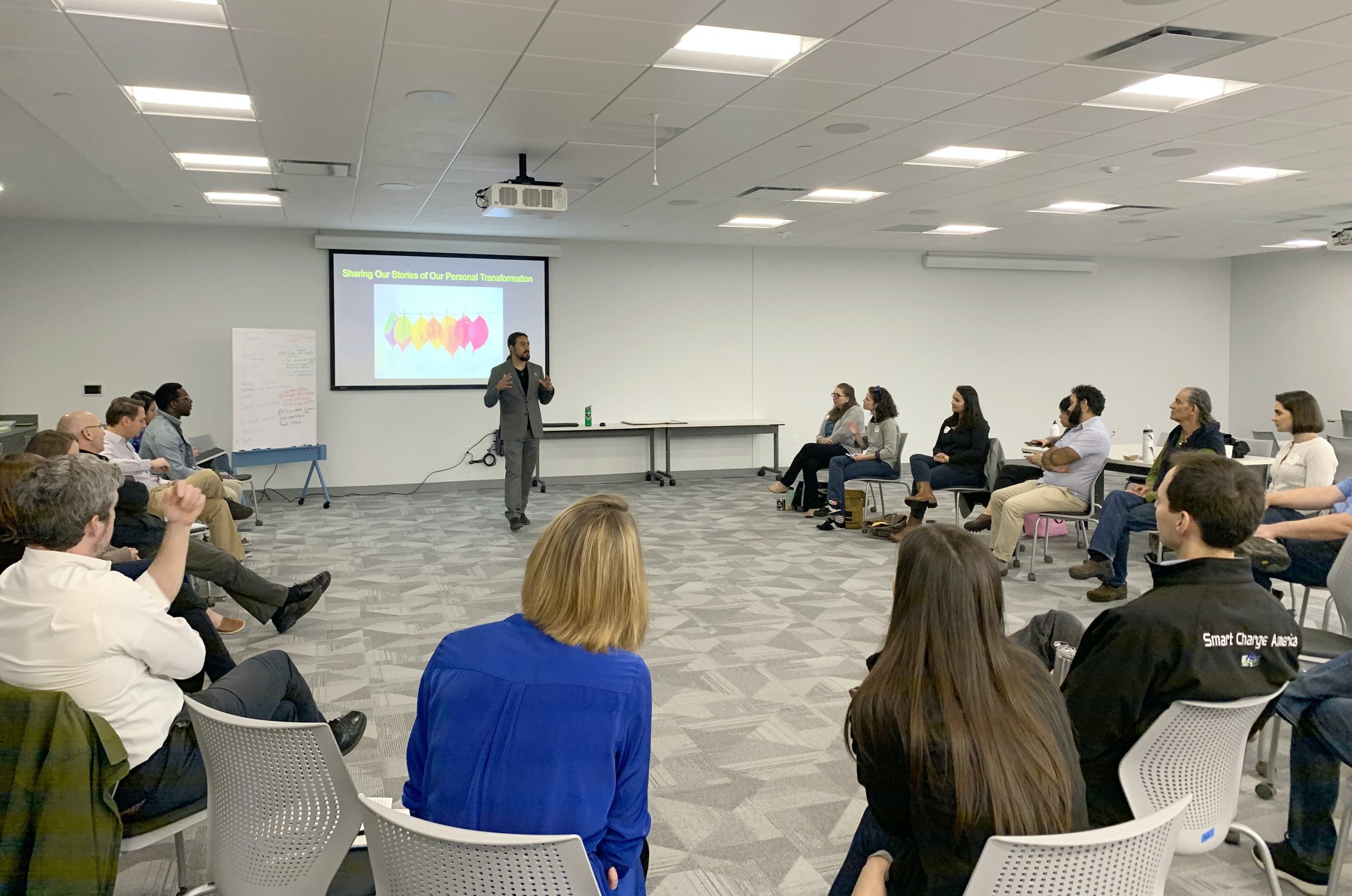 A man in a gray suit giving a presentation to a group of people seated in a classroom. The presentation slide on the wall reads 'Sharing Our Stories of Our Personal Transformation' and has a colorful graphic of speech bubbles.
