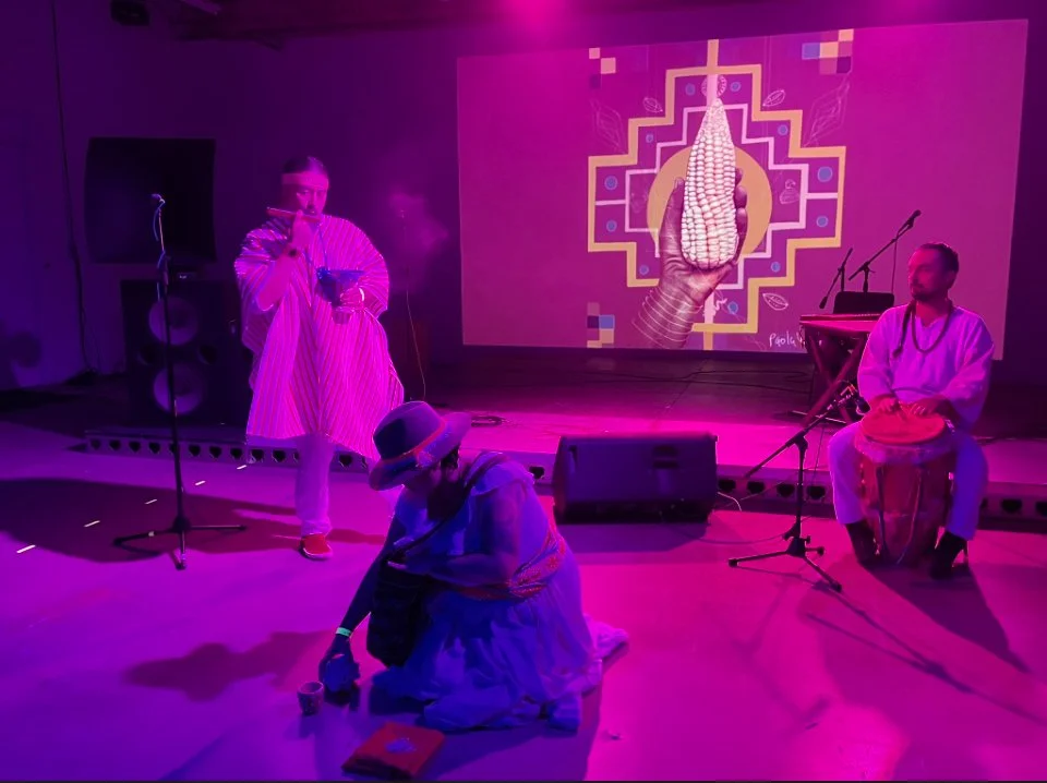Three performers on stage with a colorful digital backdrop, one holding a bowl, another sitting on the floor wearing a hat, and the third playing a drum, illuminated in pink and purple lighting.