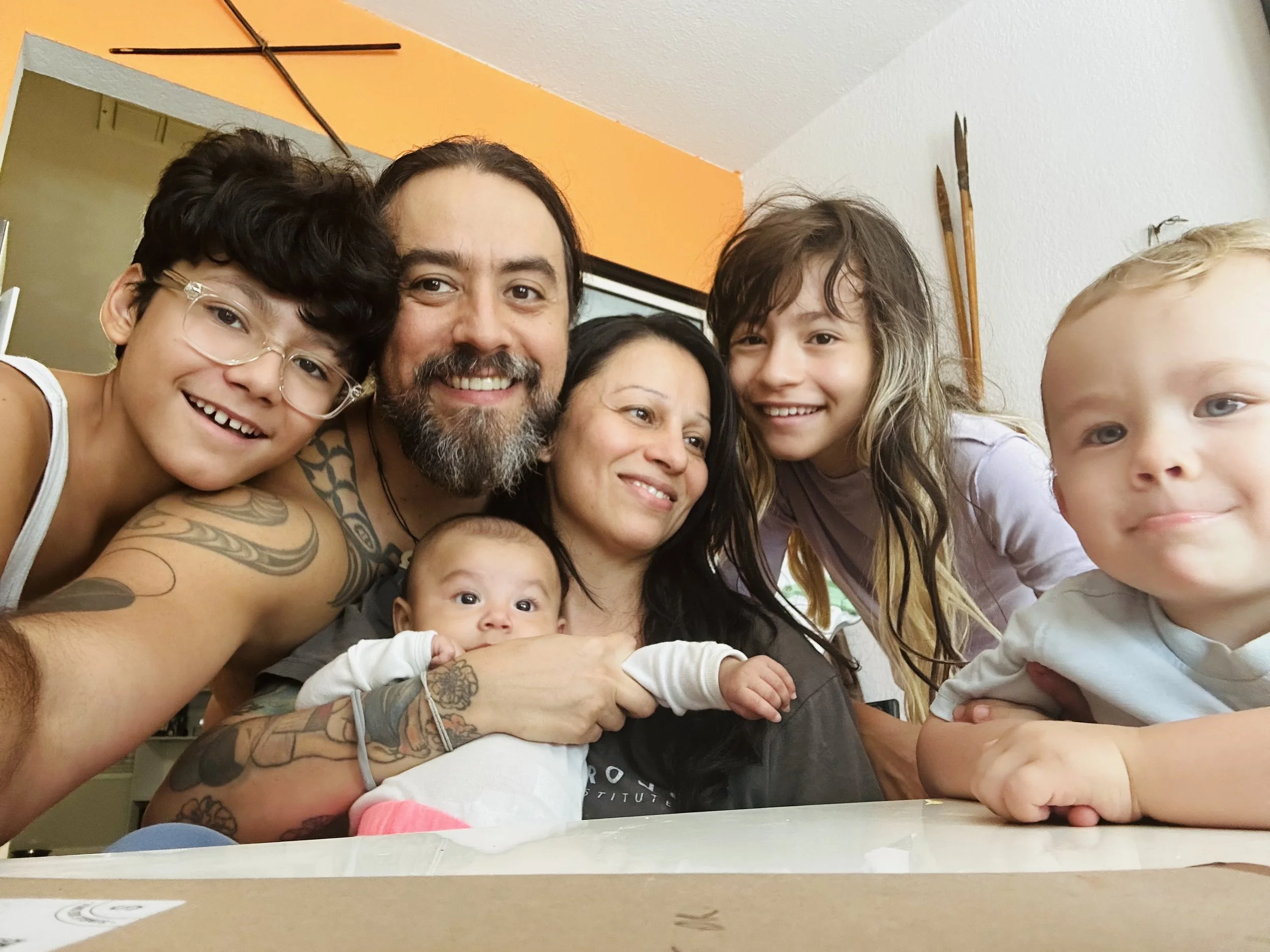 A happy family of seven taking a selfie together indoors. The group includes two adults, three children, and two babies. They are smiling and close together, with some children leaning on the adults. The background shows an orange wall with some decorative bows on it.