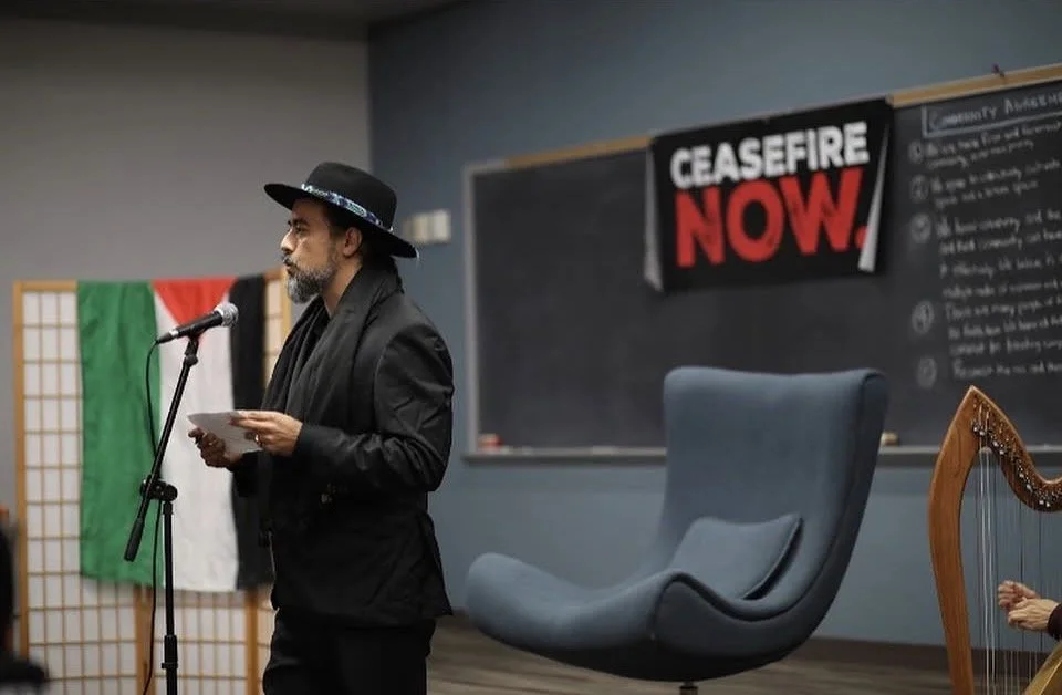 A man wearing a black suit and hat standing at a microphone, reading from a paper in a classroom setting with a 'CEASEFIRE NOW!' banner on the blackboard and a harp in the corner.