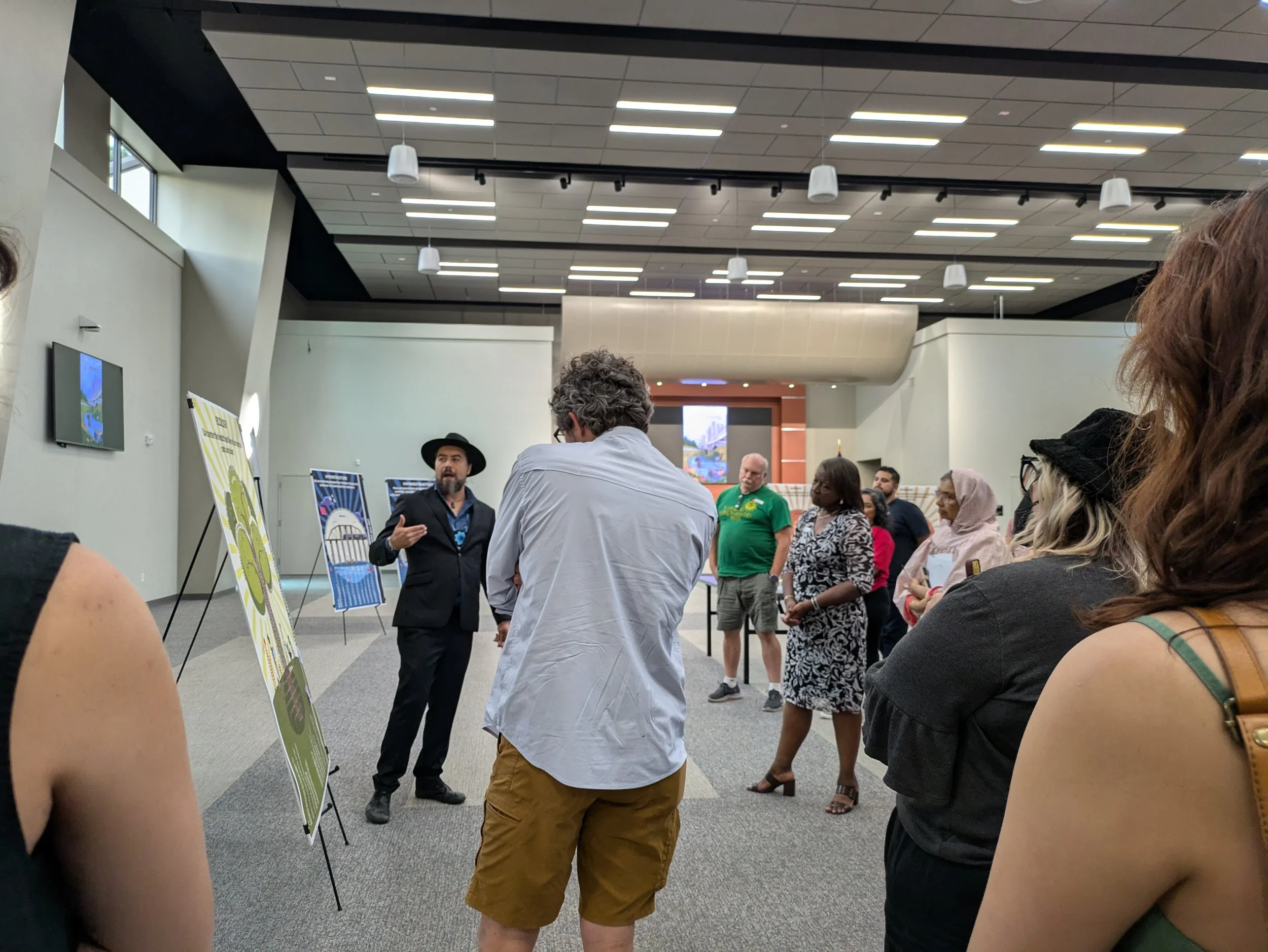 A man in a black suit and hat giving a presentation to a small group of people at an indoor event, with display posters and a screen in the background.