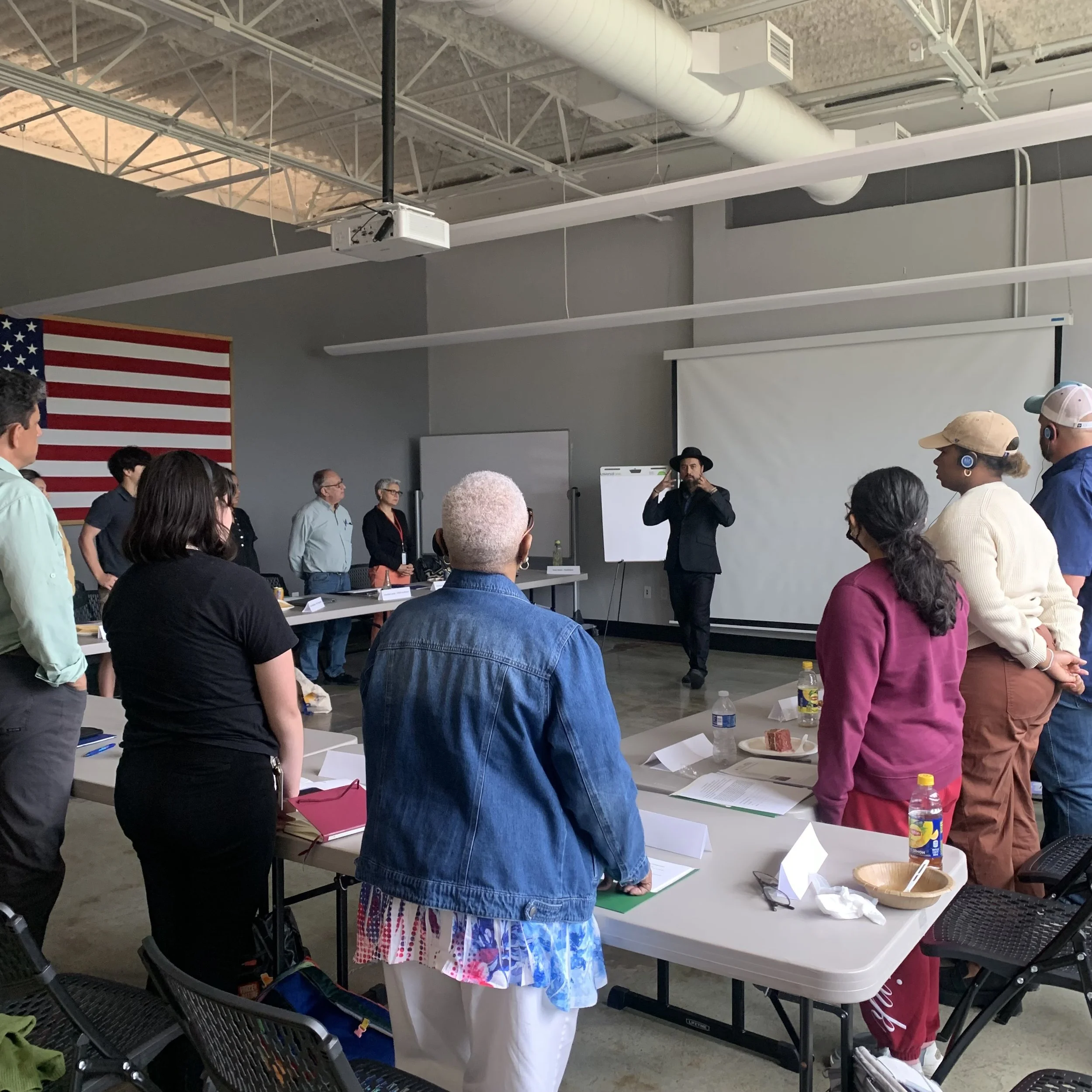 A diverse group of people standing in a room, listening to a speaker who is talking and gesturing. There is a large American flag on the wall, a whiteboard, and a projection screen in the background.