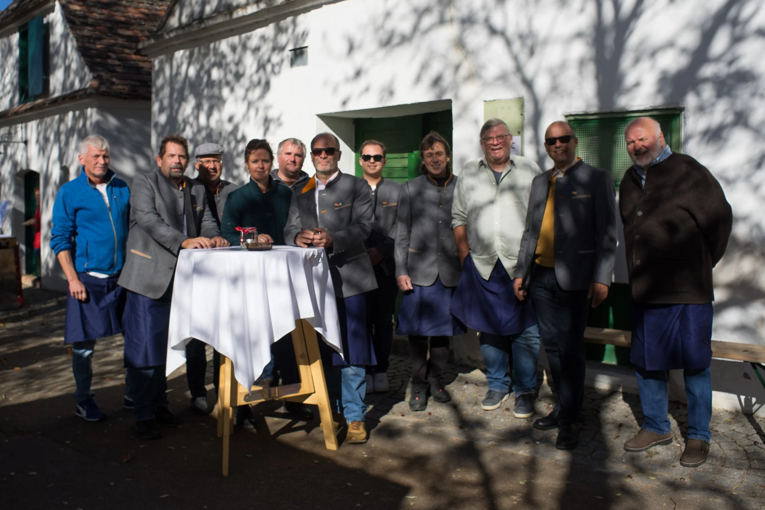 Gruppe von Menschen posiert im Schatten vor einem weißen Haus, einige tragen traditionelle Kleidung, andere moderne Kleidung, bei einer Zusammenkunft im Freien.