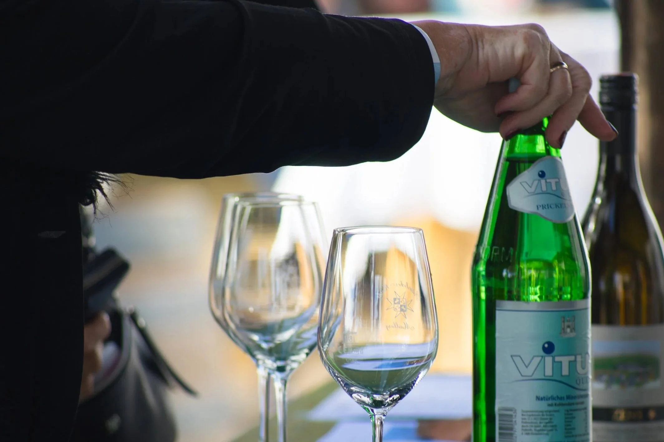 Hände öffnen eine grüne Flasche Mineralwasser während Weingläser im Vordergrund stehen.