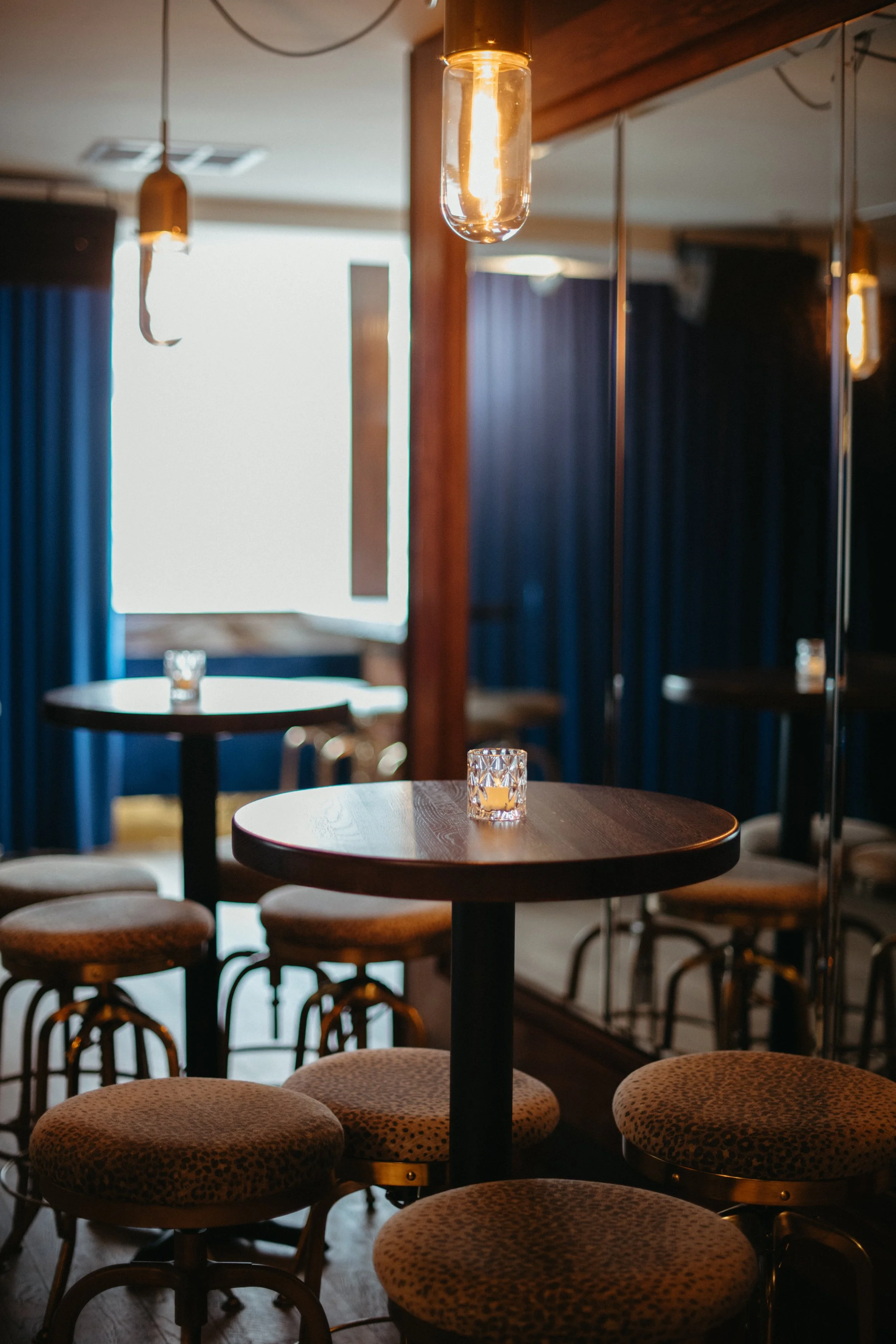 Interior of a dimly lit bar or lounge with round tables, leopard print cushioned stools, decorative hanging Edison bulbs, blue curtains, and a mirrored wall.