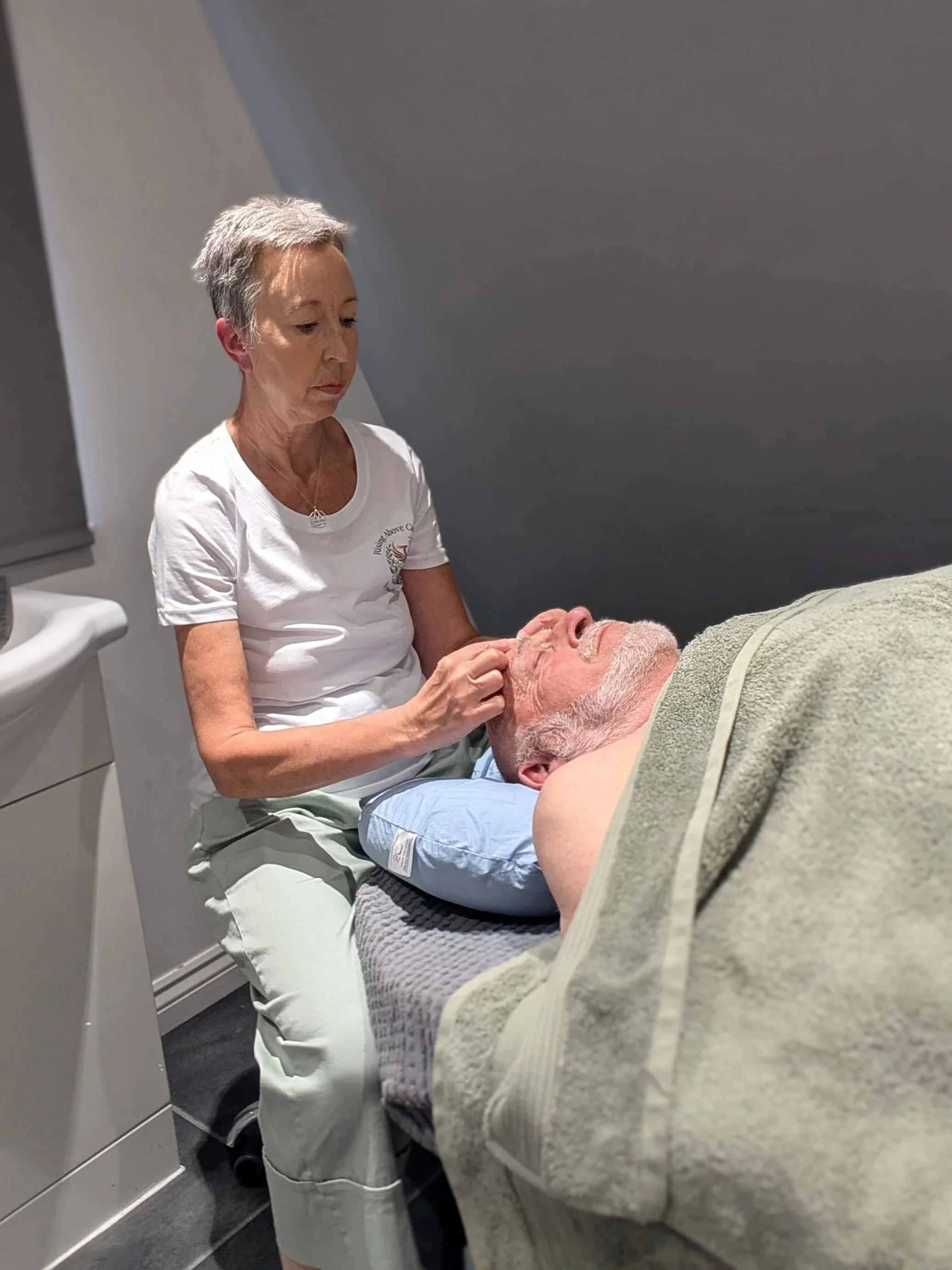 A woman giving a facial treatment to a man lying on a treatment bed in a spa or clinic setting.
