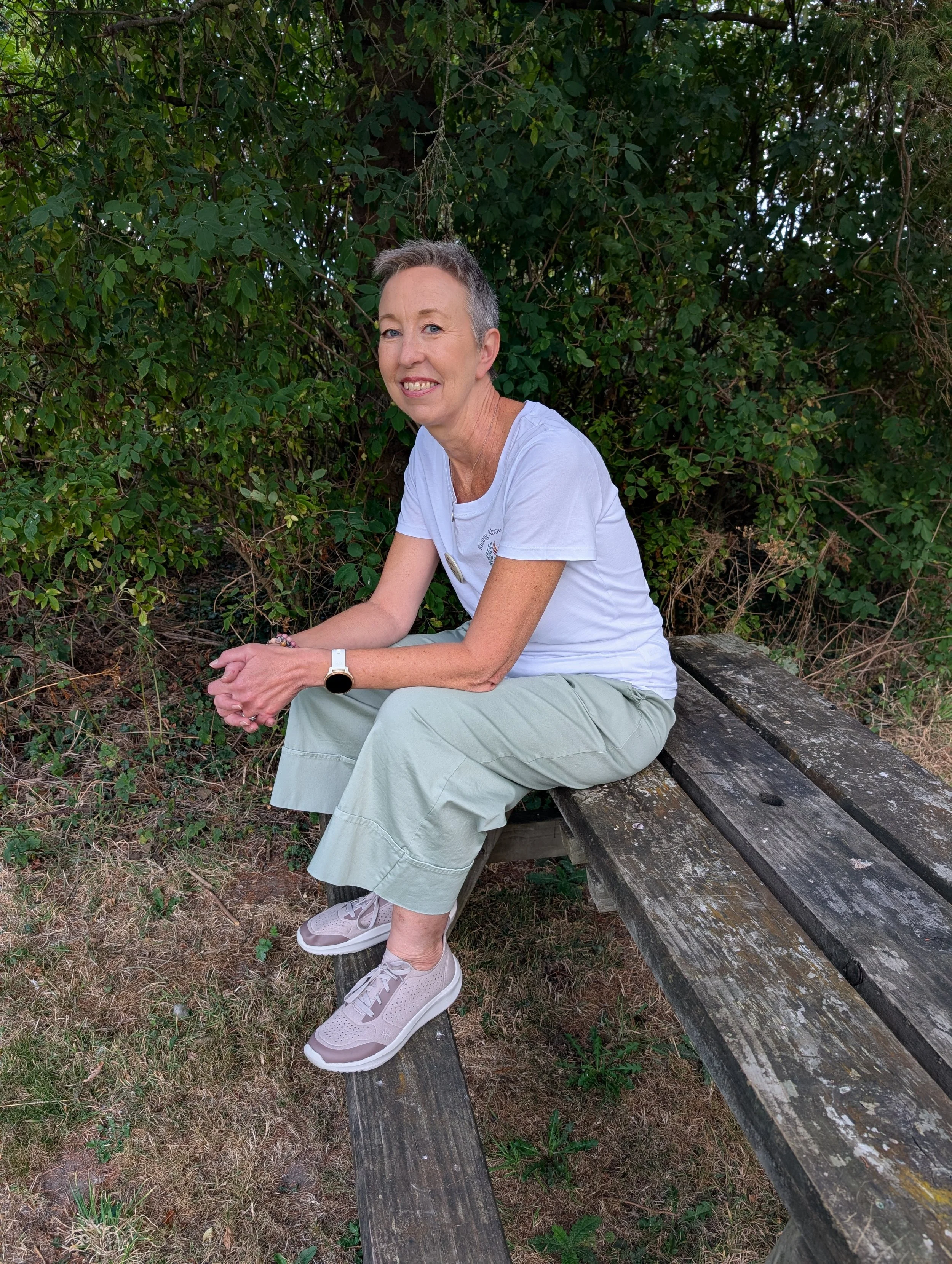 A woman with short gray hair is sitting on a weathered wooden bench in front of a thick green bush. She is smiling and wearing a white t-shirt, light-colored pants, and pink sneakers. She has a smartwatch on her left wrist and a beaded bracelet on her right wrist.