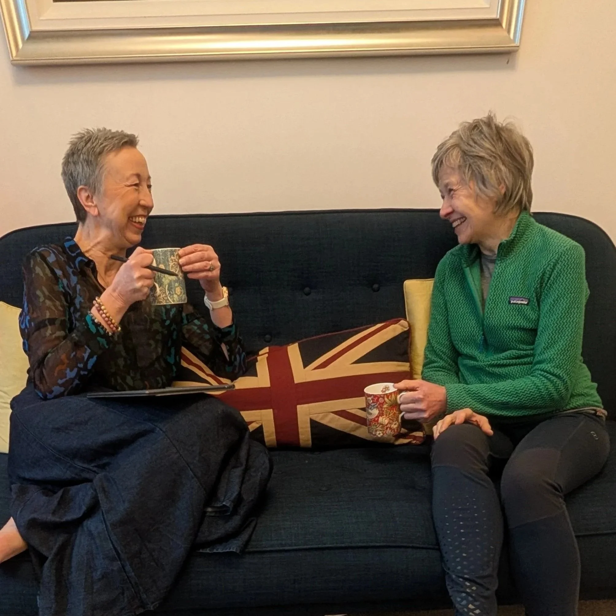 Two women are sitting on a black cushioned bench, smiling and interacting with each other in a cozy indoor setting. The woman on the left has short gray hair, is wearing a multicolored patterned blouse, and is holding a cup while using chopsticks. The woman on the right has short blonde hair, is wearing a bright green sweater, and holds a Christmas-themed mug. The background has a beige wall with a large gold picture frame and a cushion with a Union Jack design on the bench.