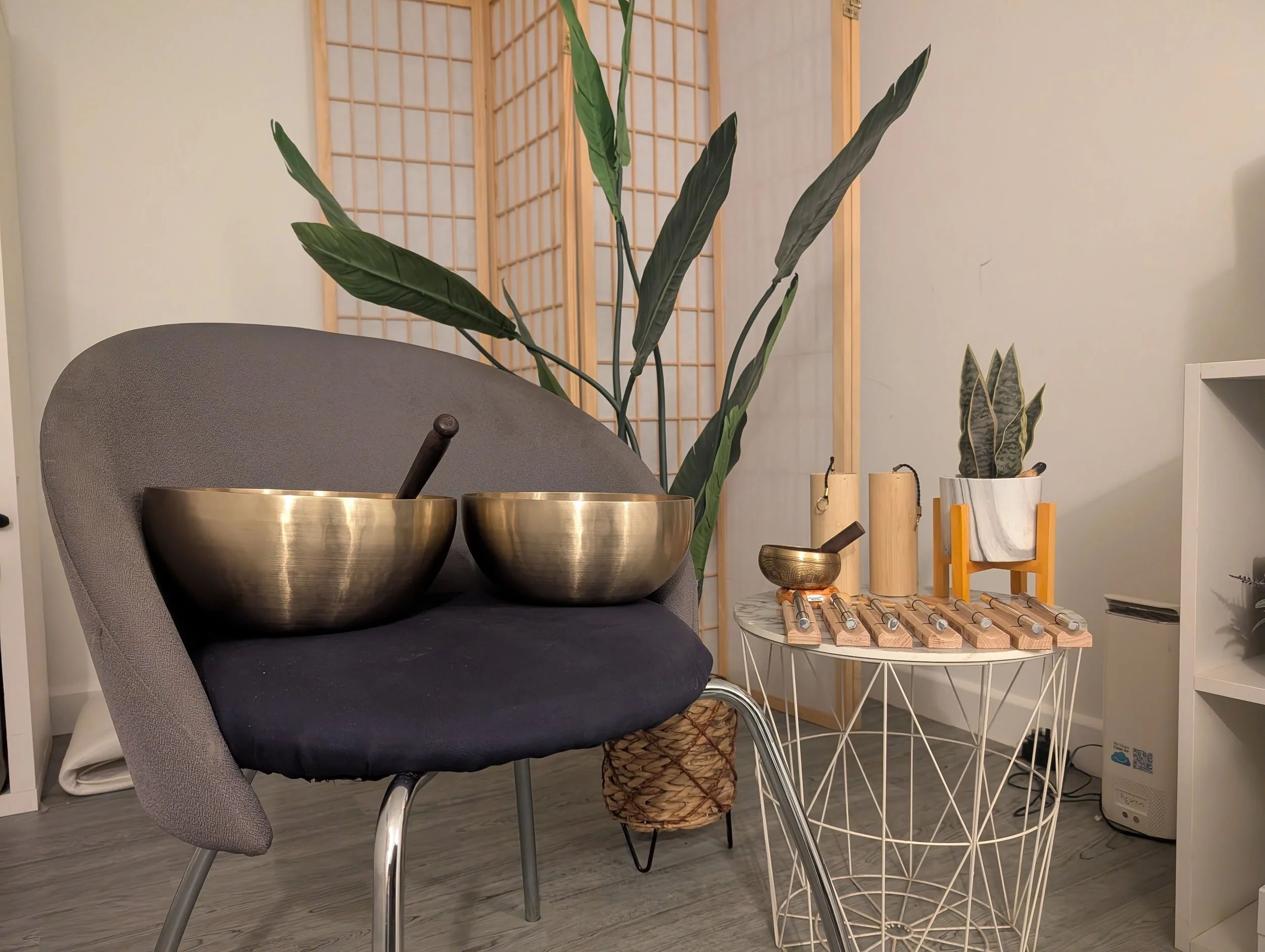 Interior room with a gray chair, a round side table, tall indoor plants, a wooden room divider, and decorative objects including metal bowls and a rattle.