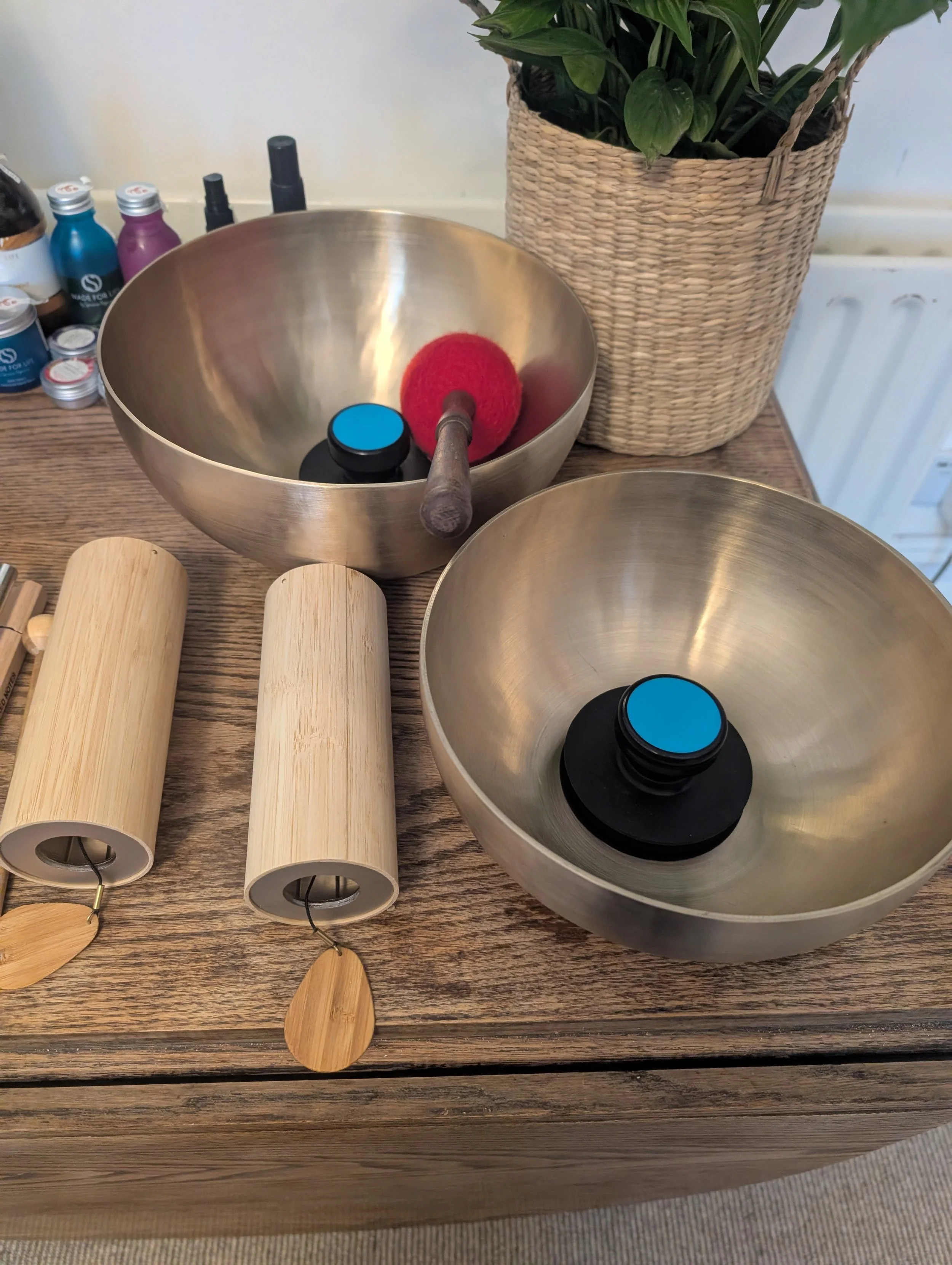 Two handpan musical instruments with wooden mallets on a wooden table, a potted plant in a woven basket, and bottles of paint alongside spray bottles in the background.