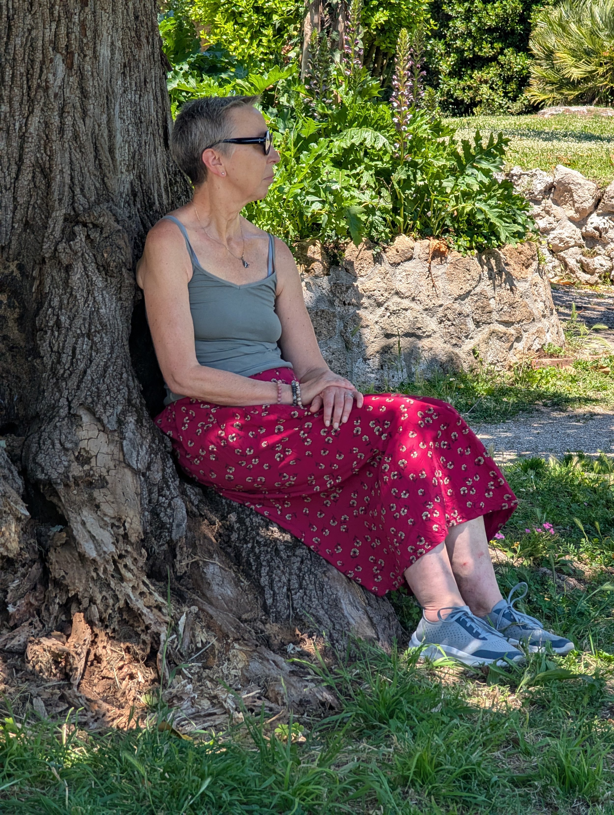 A woman with short gray hair, wearing sunglasses, a gray tank top, and a red patterned skirt, sitting against a large tree trunk in a lush, sunny garden with rocks and greenery.